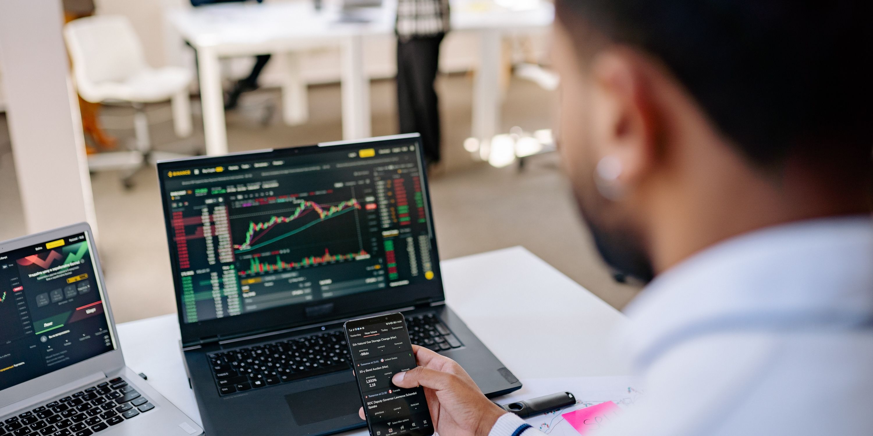 A man flips through an app on his phone, while perusing graphs on a laptop behind the phone.