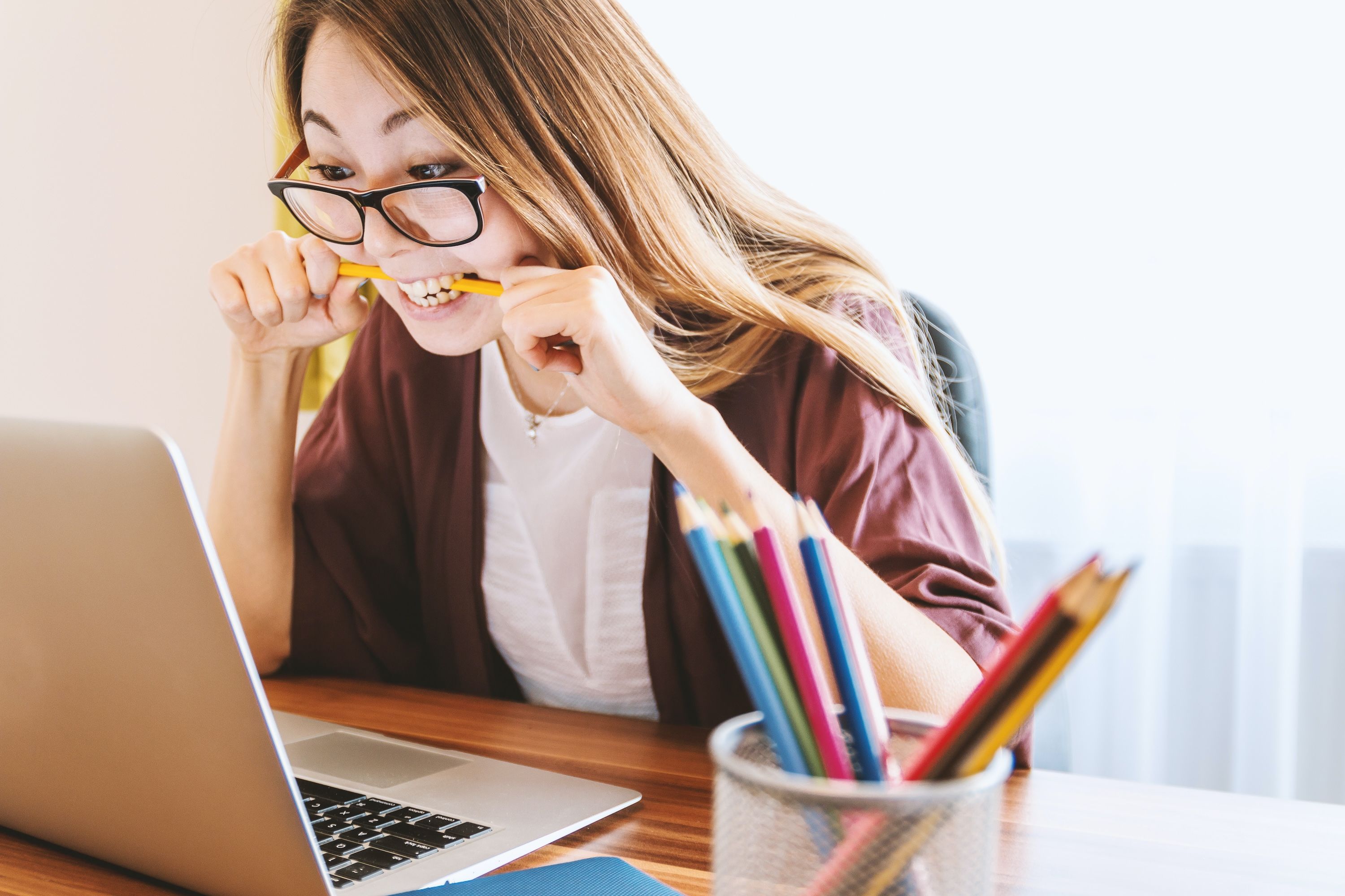 A woman wearing glasses bites a pencil while working on finances on her laptop.