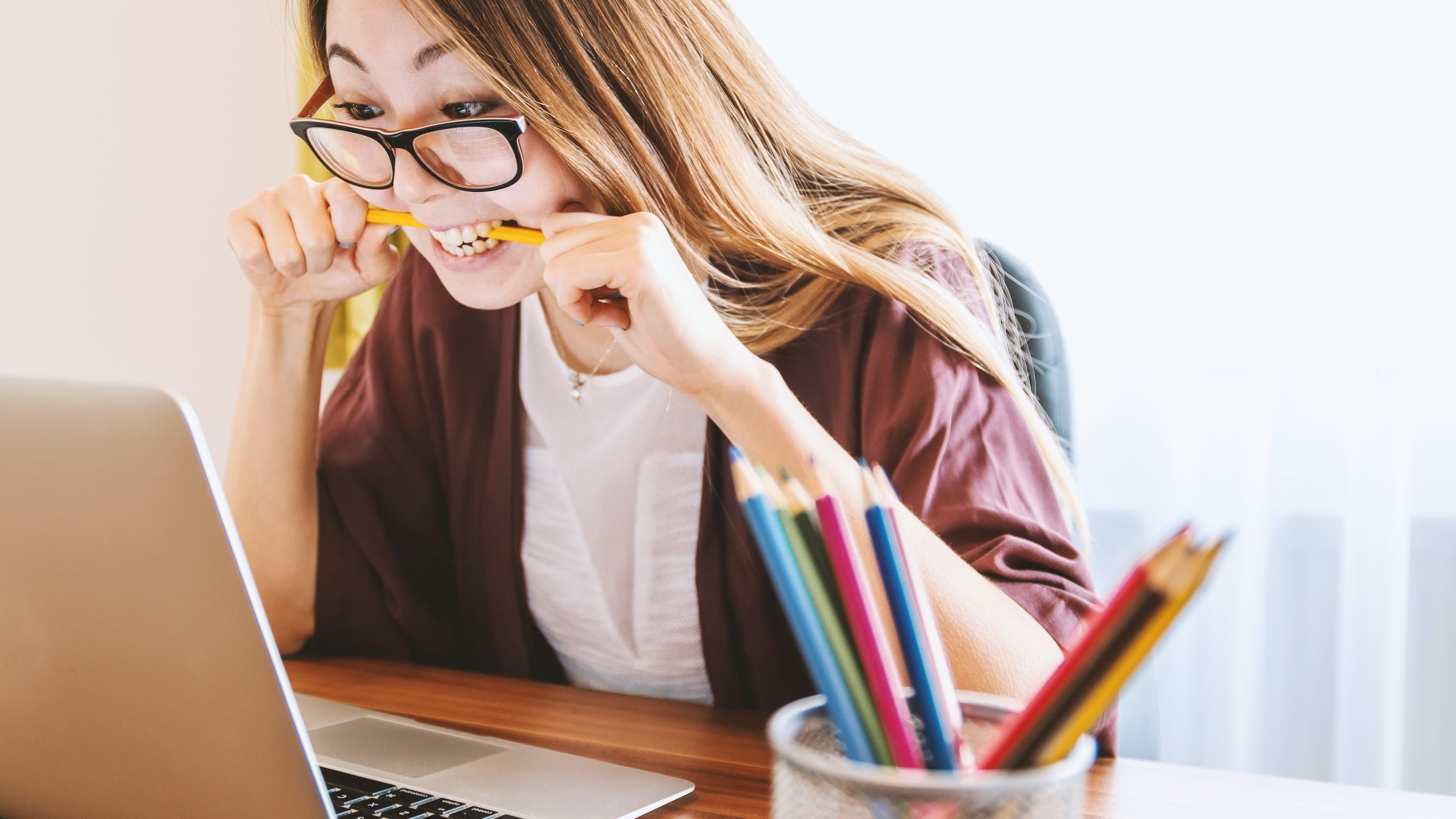 A woman wearing glasses bites a pencil while working on finances on her laptop.