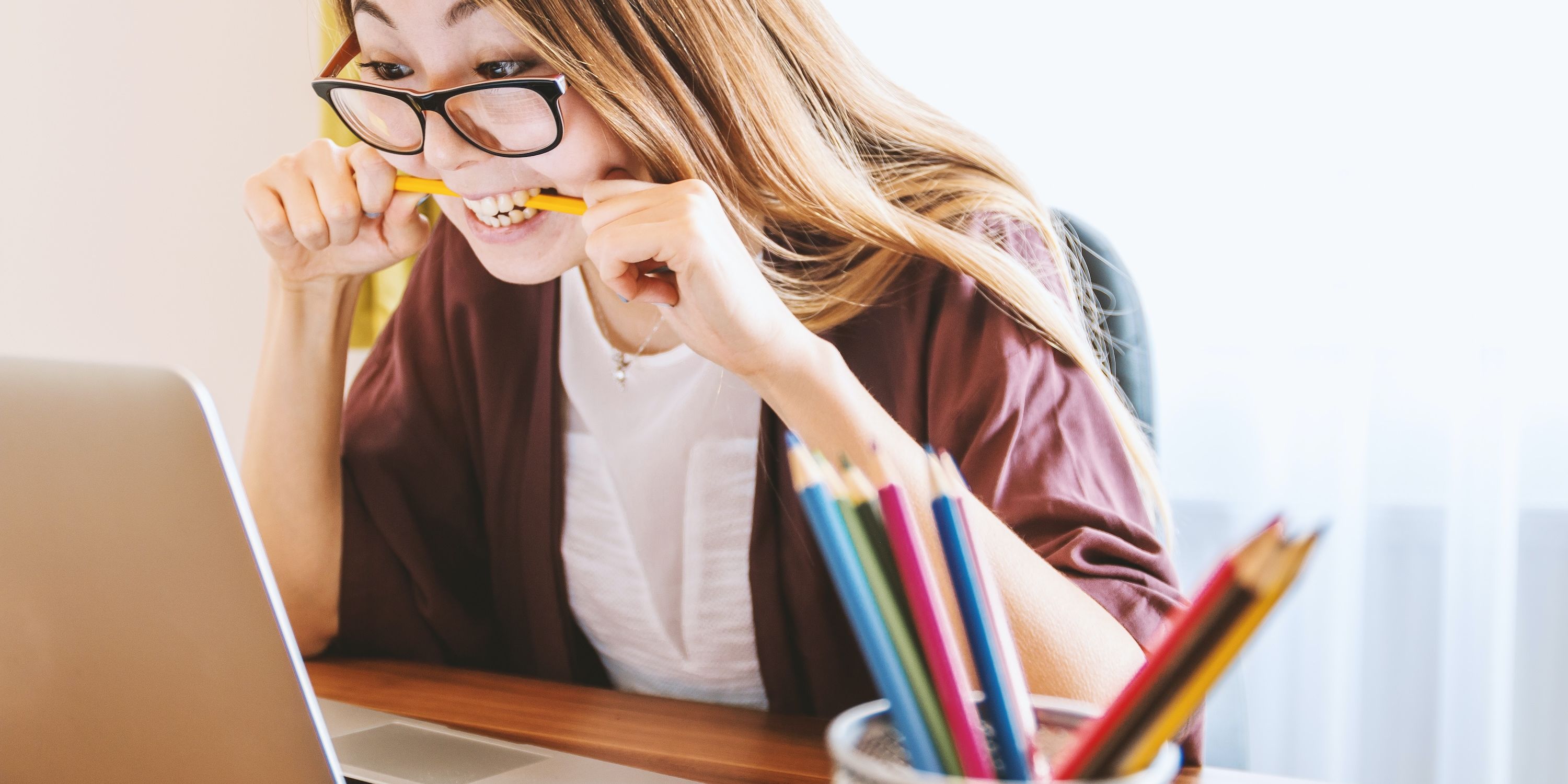 A woman wearing glasses bites a pencil while working on finances on her laptop.