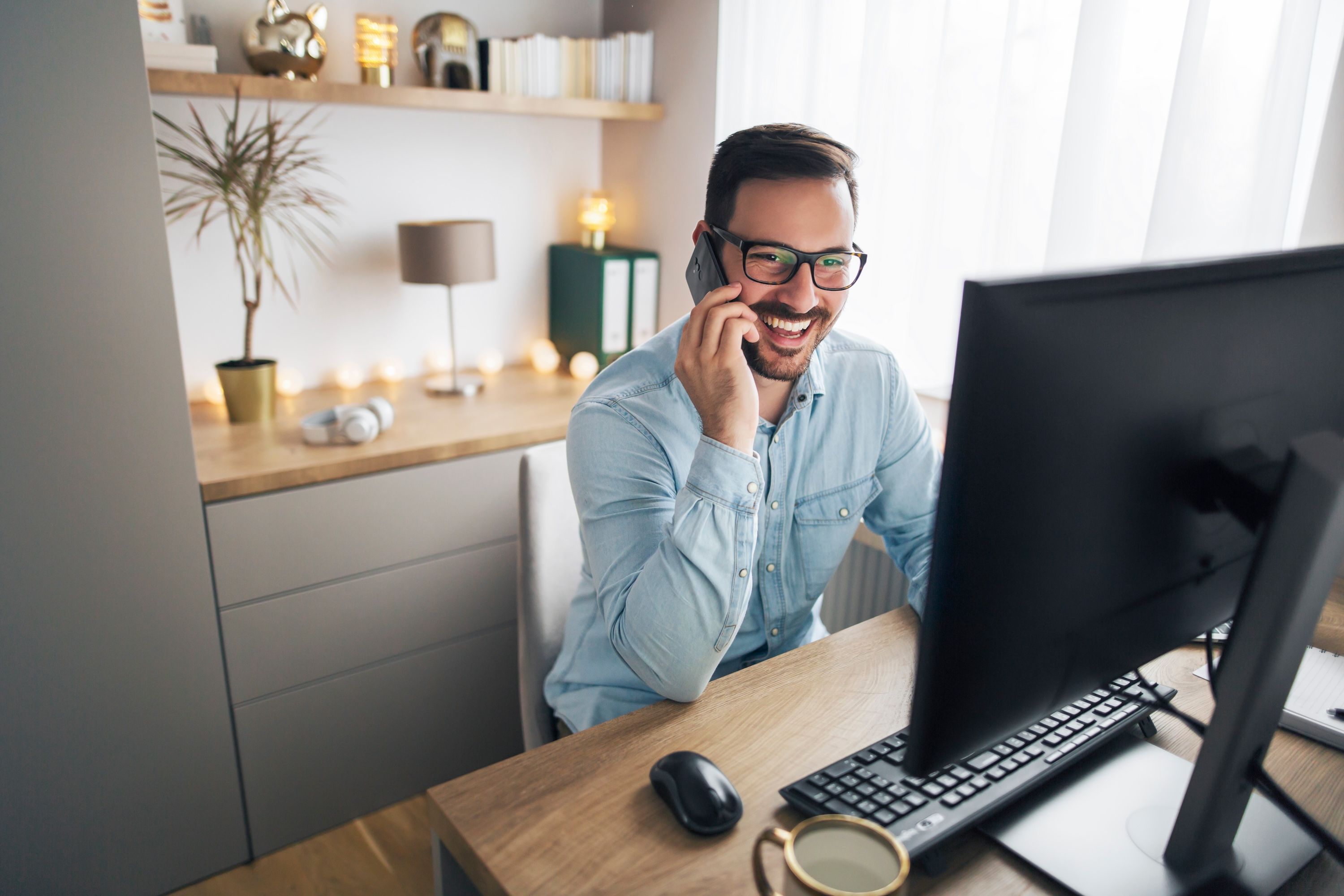 A man wearing glasses and a blue shirt smiles and laughs on the phone while looking at a computer screen.