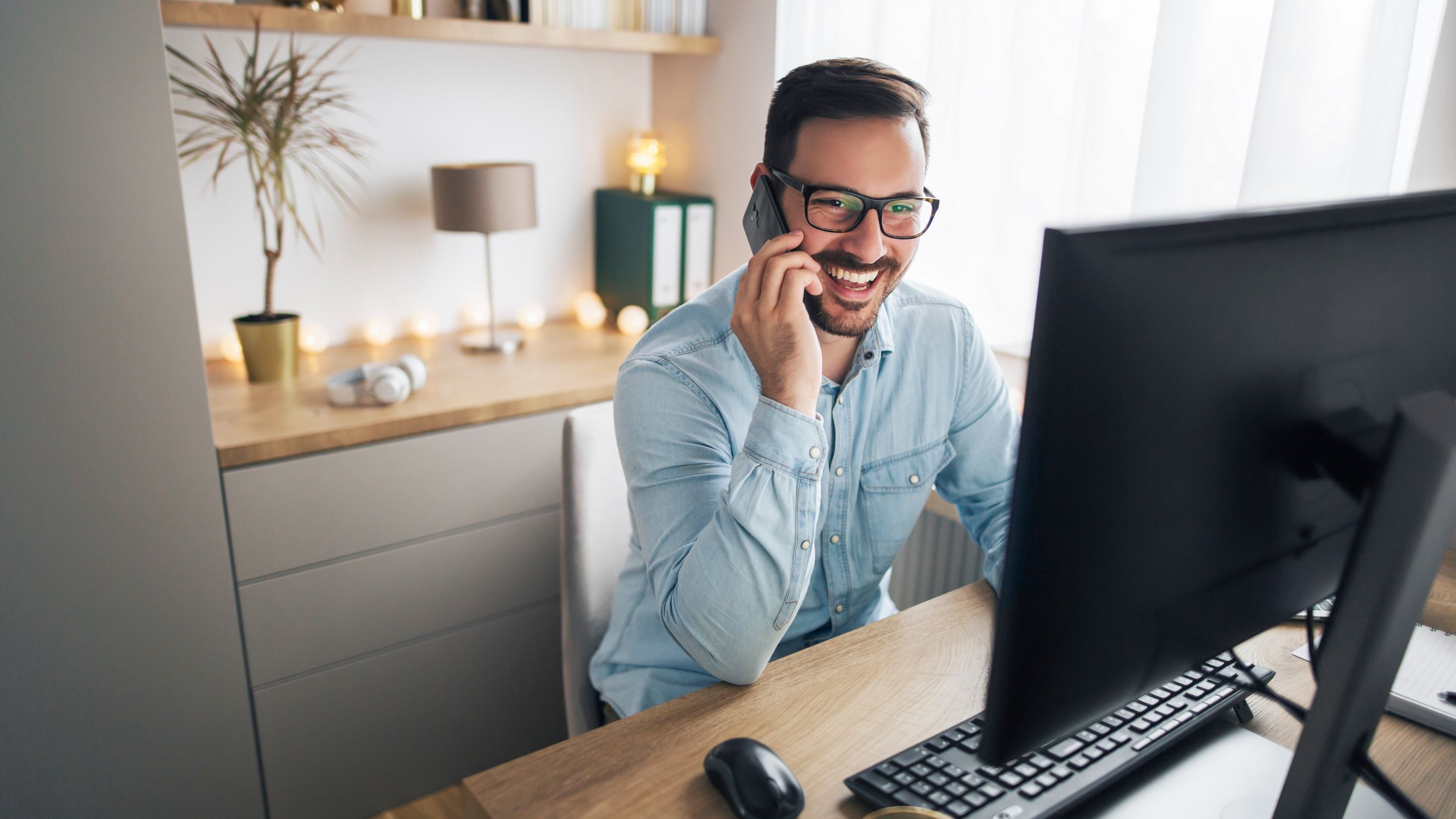 A man wearing glasses and a blue shirt smiles and laughs on the phone while looking at a computer screen.
