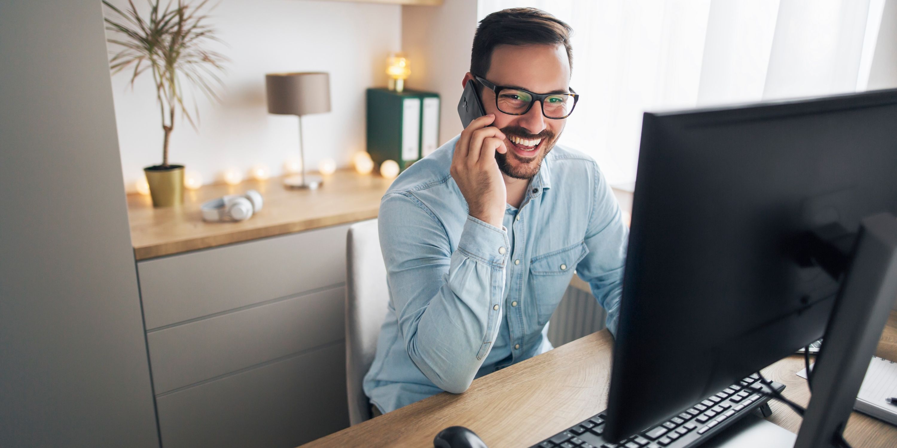A man wearing glasses and a blue shirt smiles and laughs on the phone while looking at a computer screen.