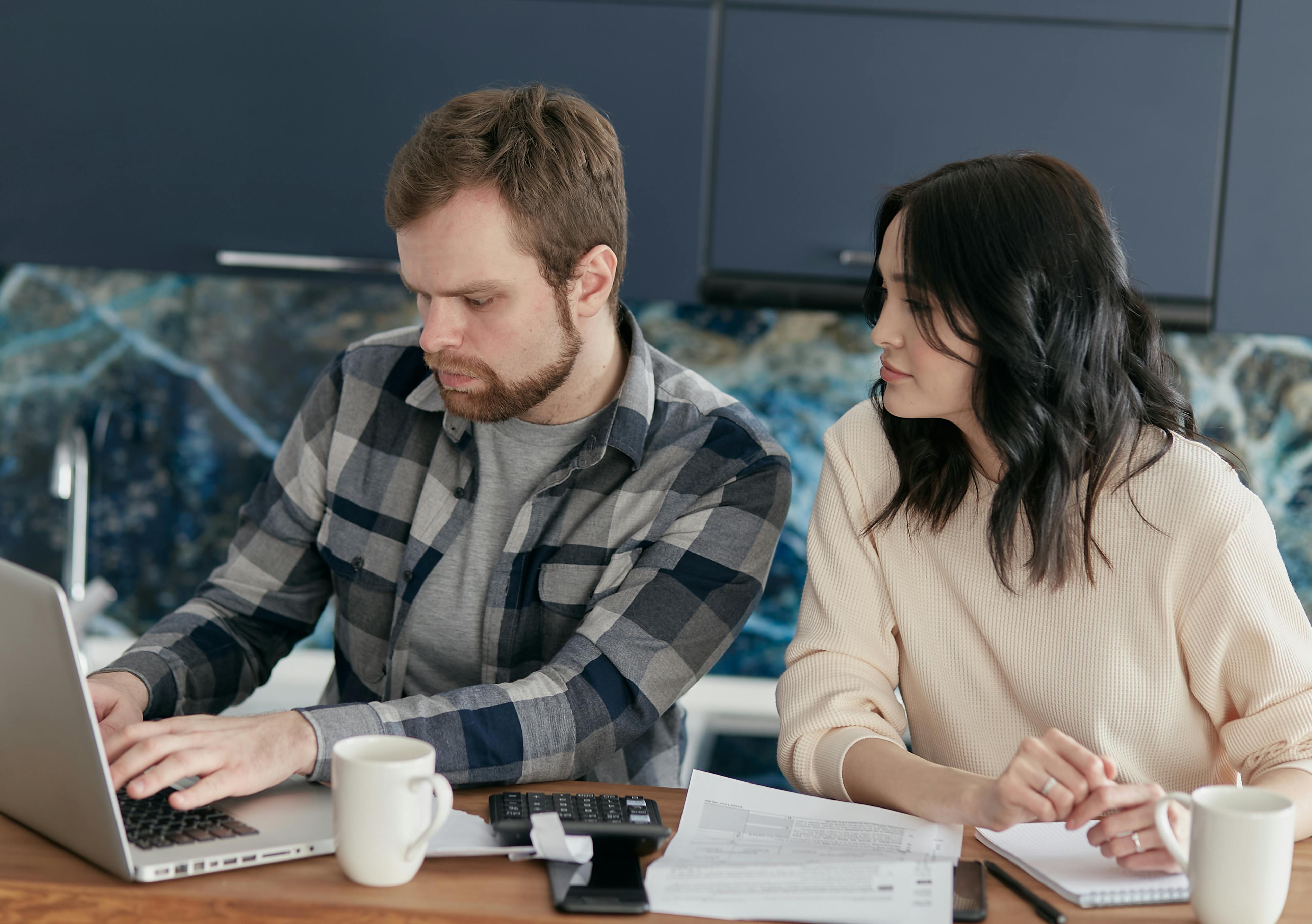 A concerned-looking couple works together on finances with a sprawl of coffee, calculators and notebooks surrounding them while they work on a laptop.