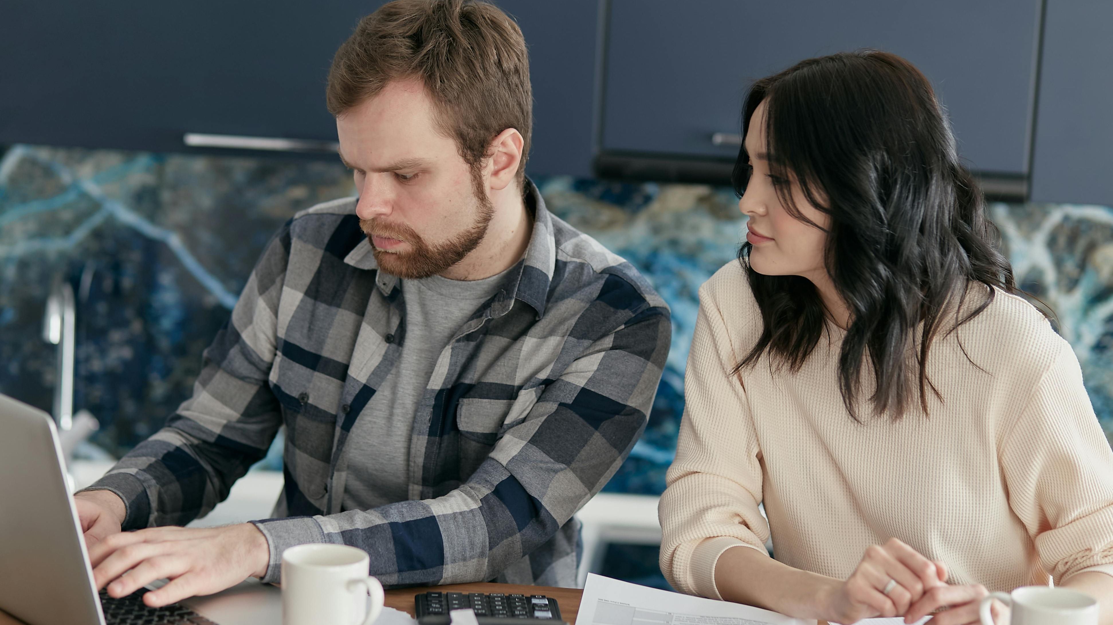 A concerned-looking couple works together on finances with a sprawl of coffee, calculators and notebooks surrounding them while they work on a laptop.