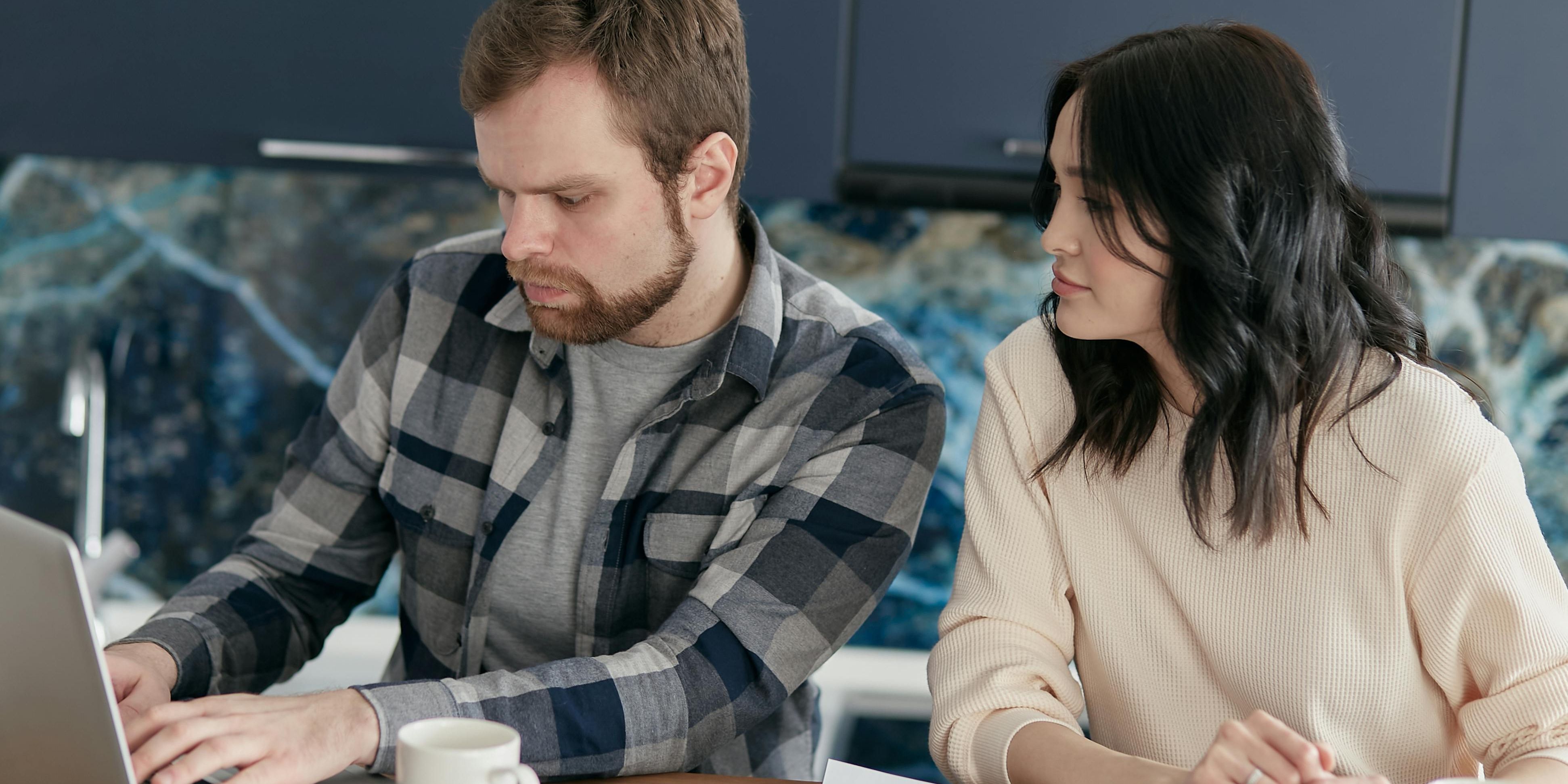 A concerned-looking couple works together on finances with a sprawl of coffee, calculators and notebooks surrounding them while they work on a laptop.