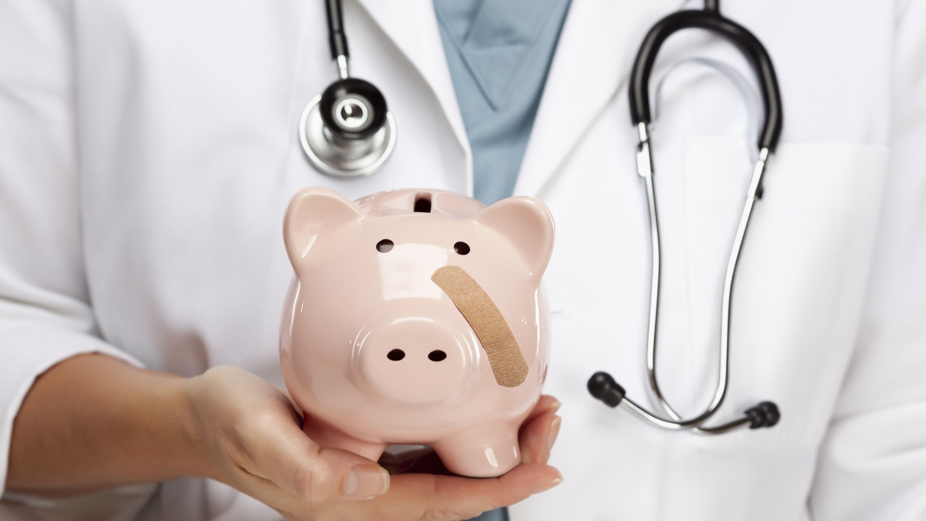 A doctor holds up a piggy bank with a band-aid on its cheek.