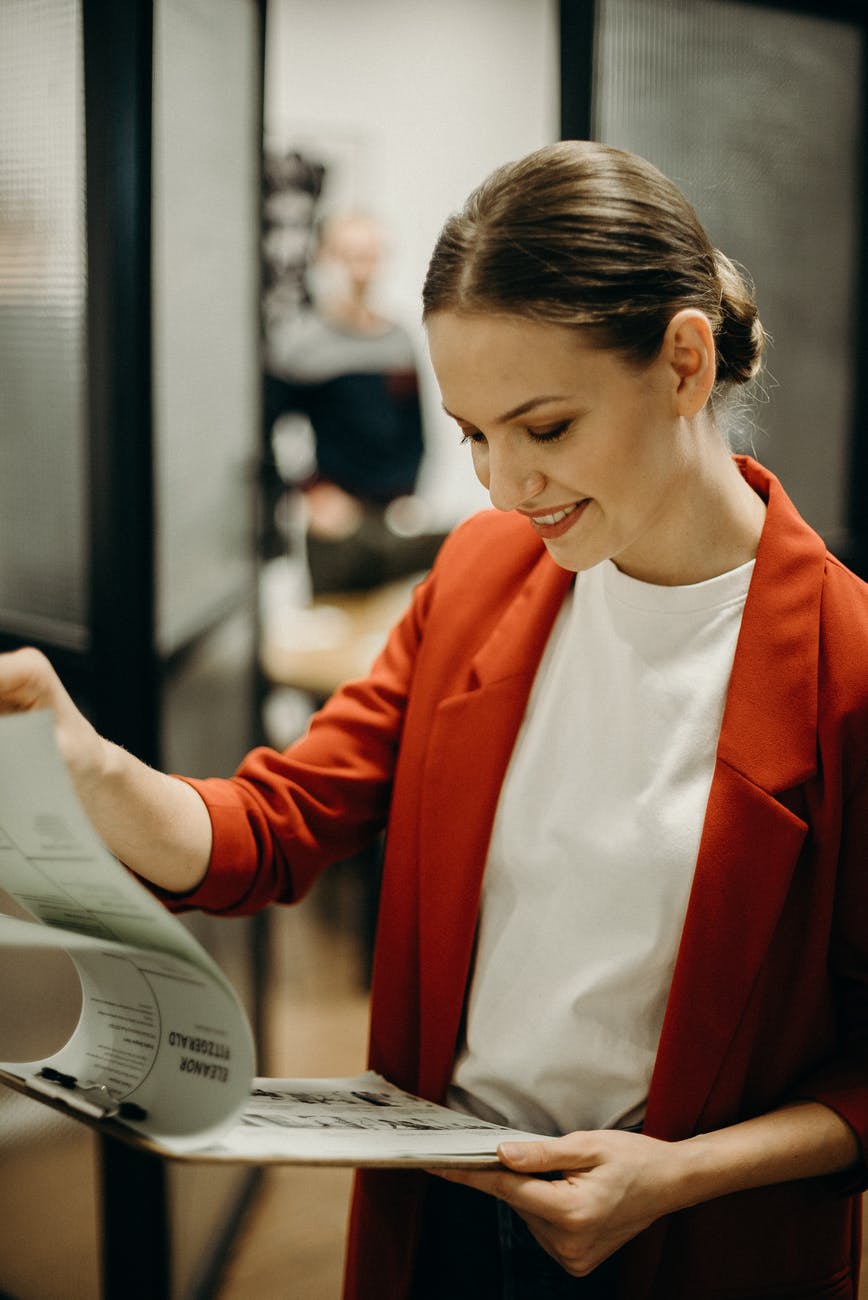 A woman wearing a red jacket peruses papers on a clipboard while at the office.