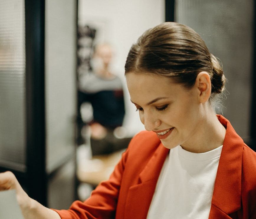 A woman wearing a red jacket peruses papers on a clipboard while at the office.