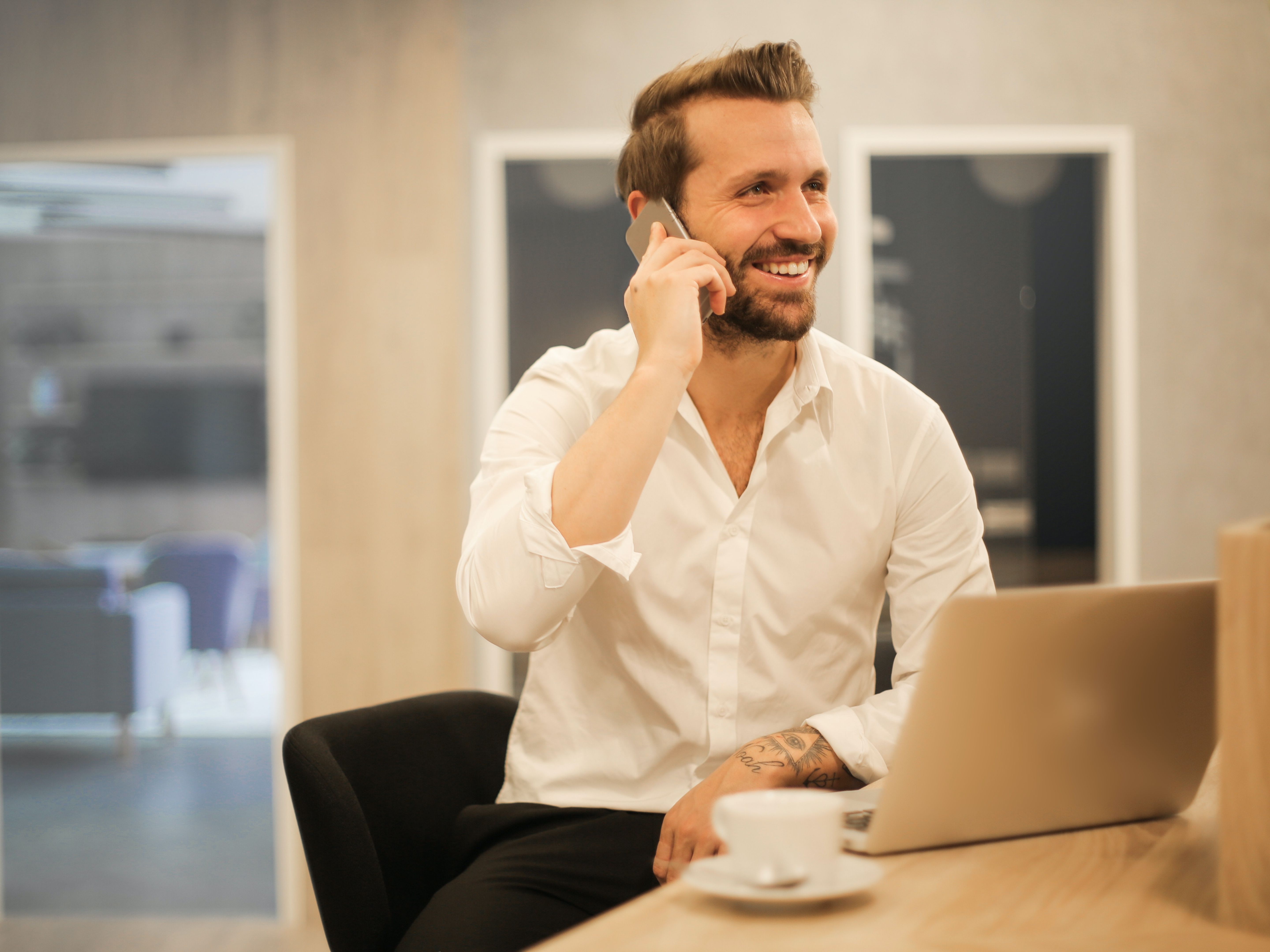 A smiling man talks on a phone while sitting at a desk near a laptop and coffee.