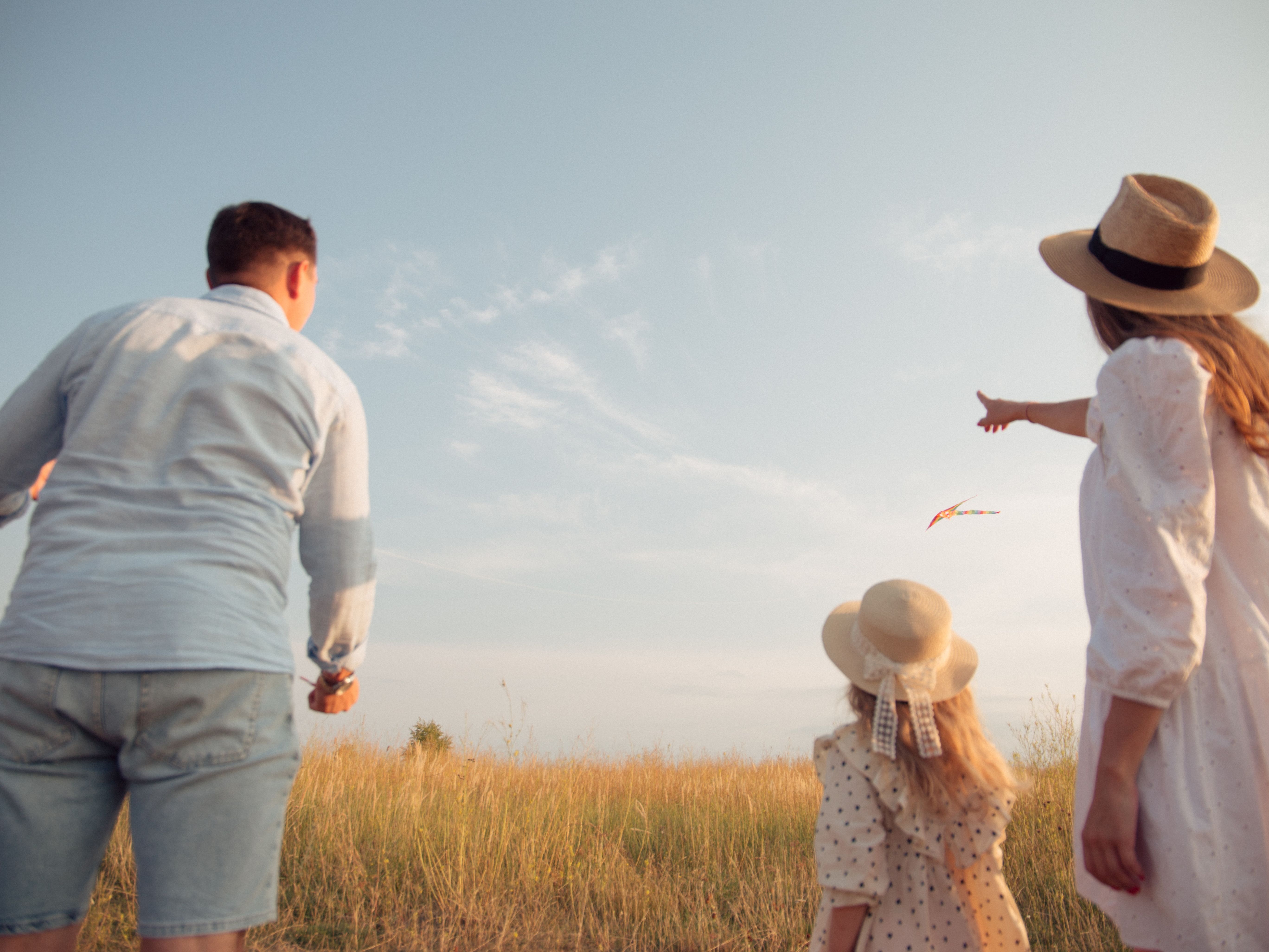 Two parents run after a kite with a child, while the mother points at a cloud.