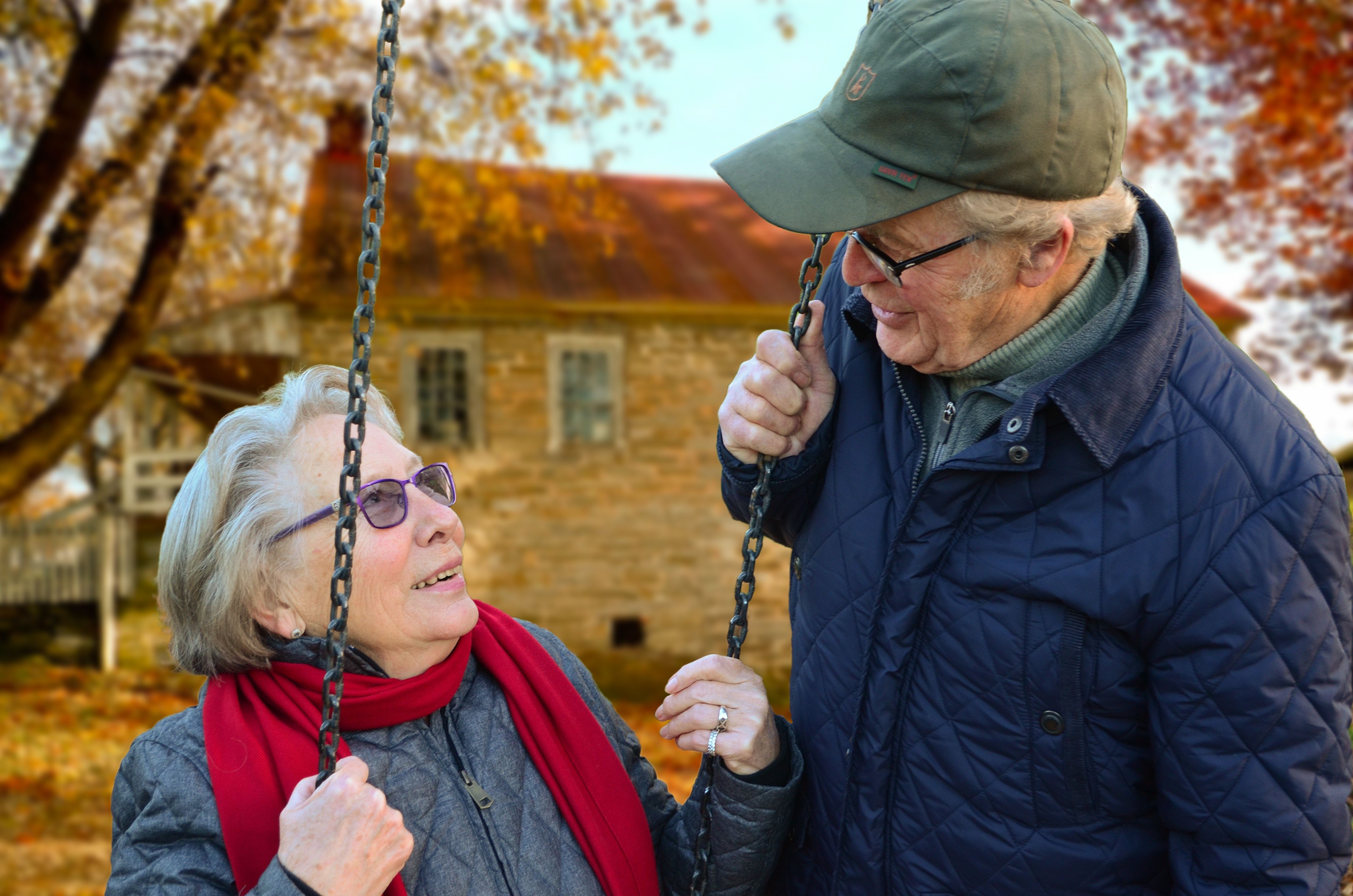 An elderly couple smile at each other while the woman sits on a swing during the autumn.