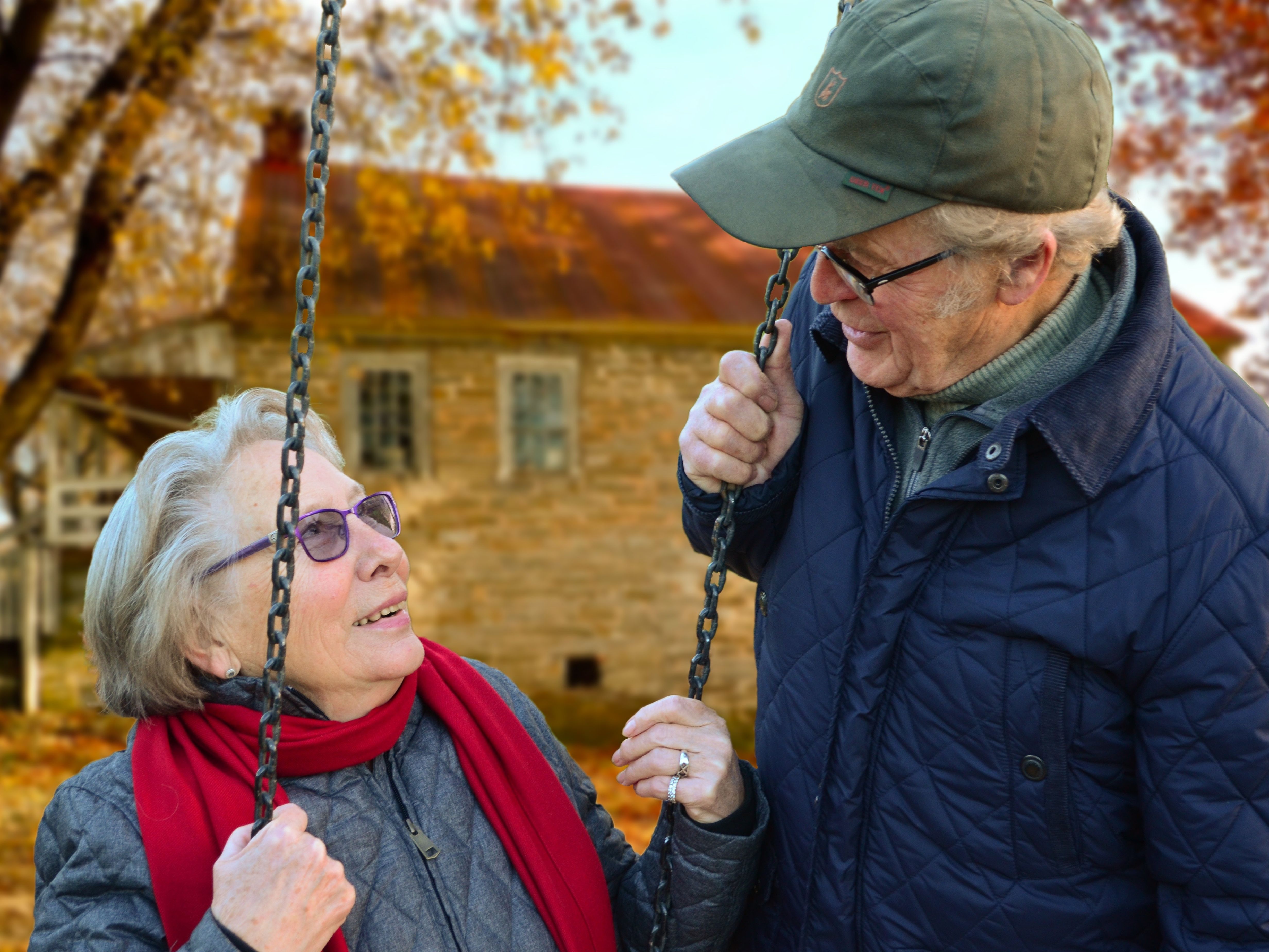 An elderly couple smile at each other while the woman sits on a swing during the autumn.