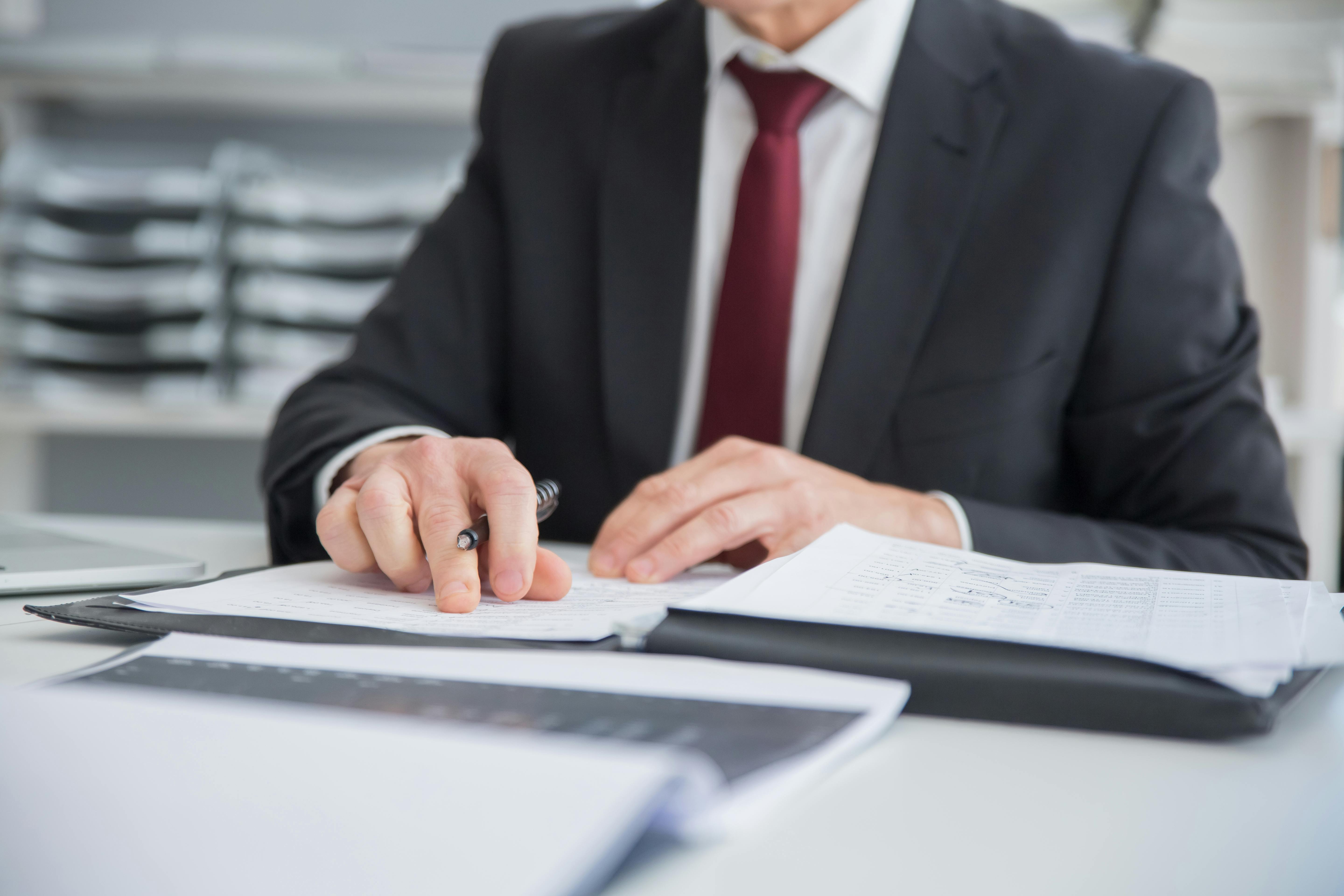 A person in a suit with a red tie works through some papers on a white desk.