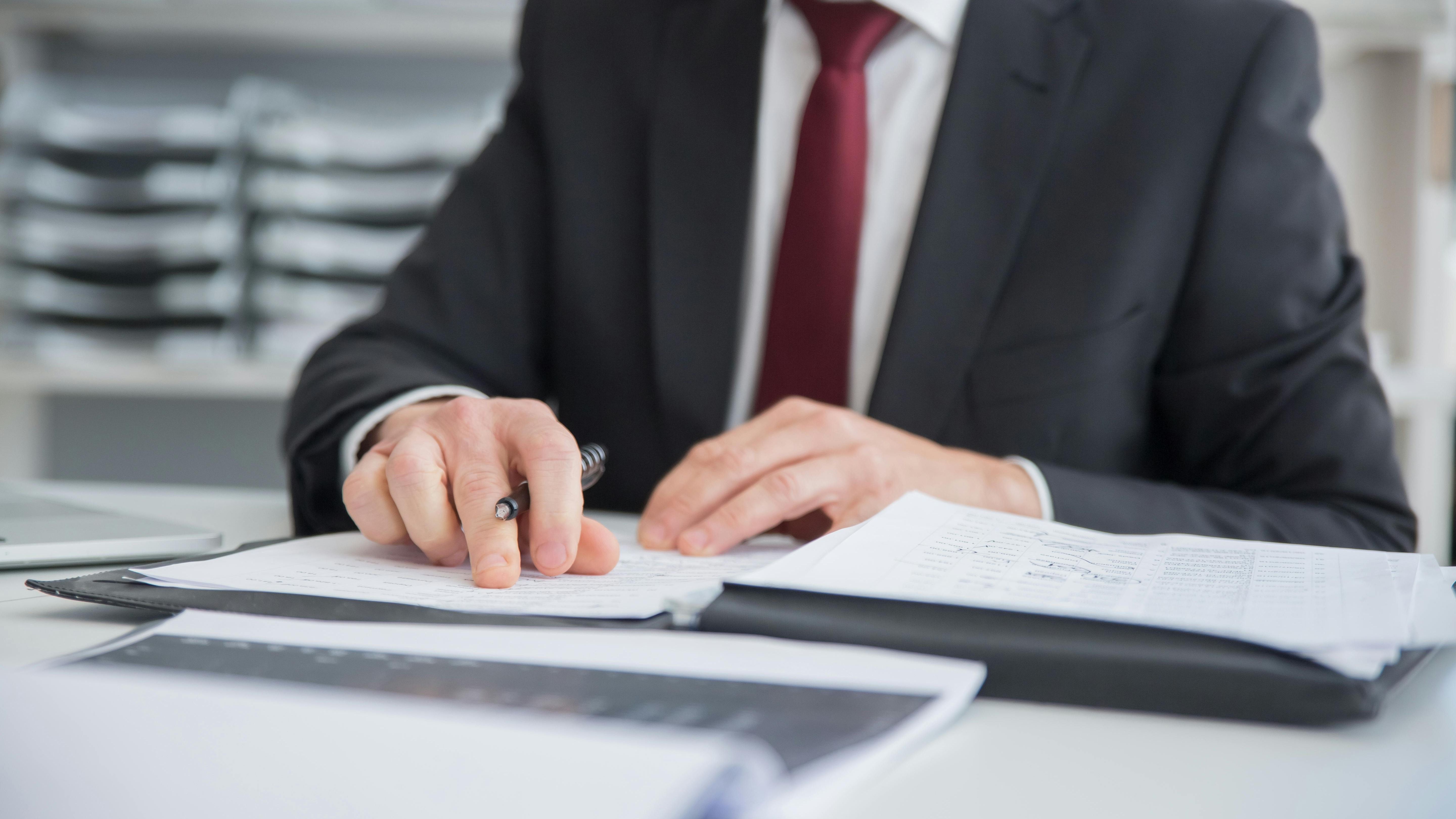 A person in a suit with a red tie works through some papers on a white desk.