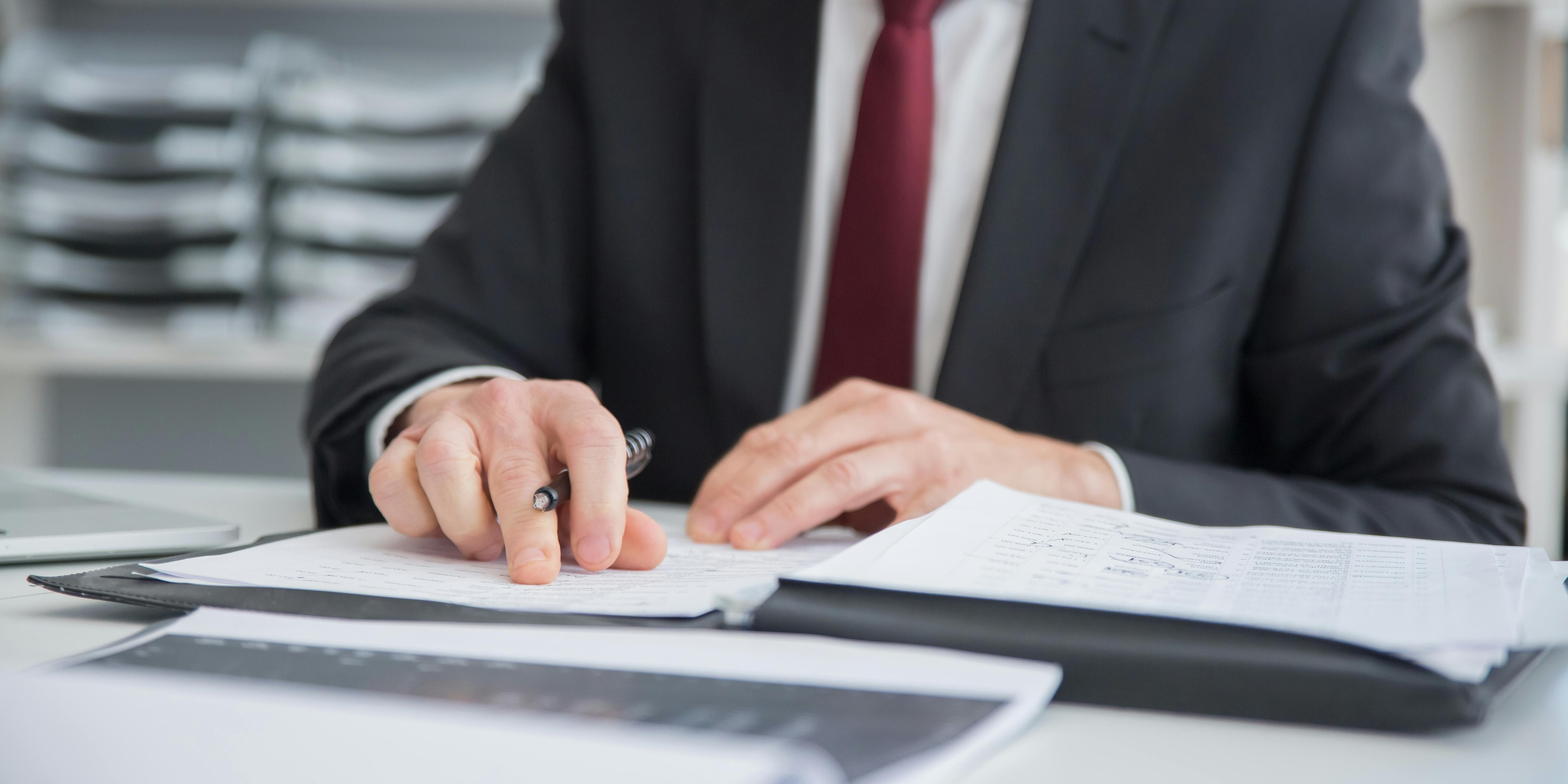 A person in a suit with a red tie works through some papers on a white desk.