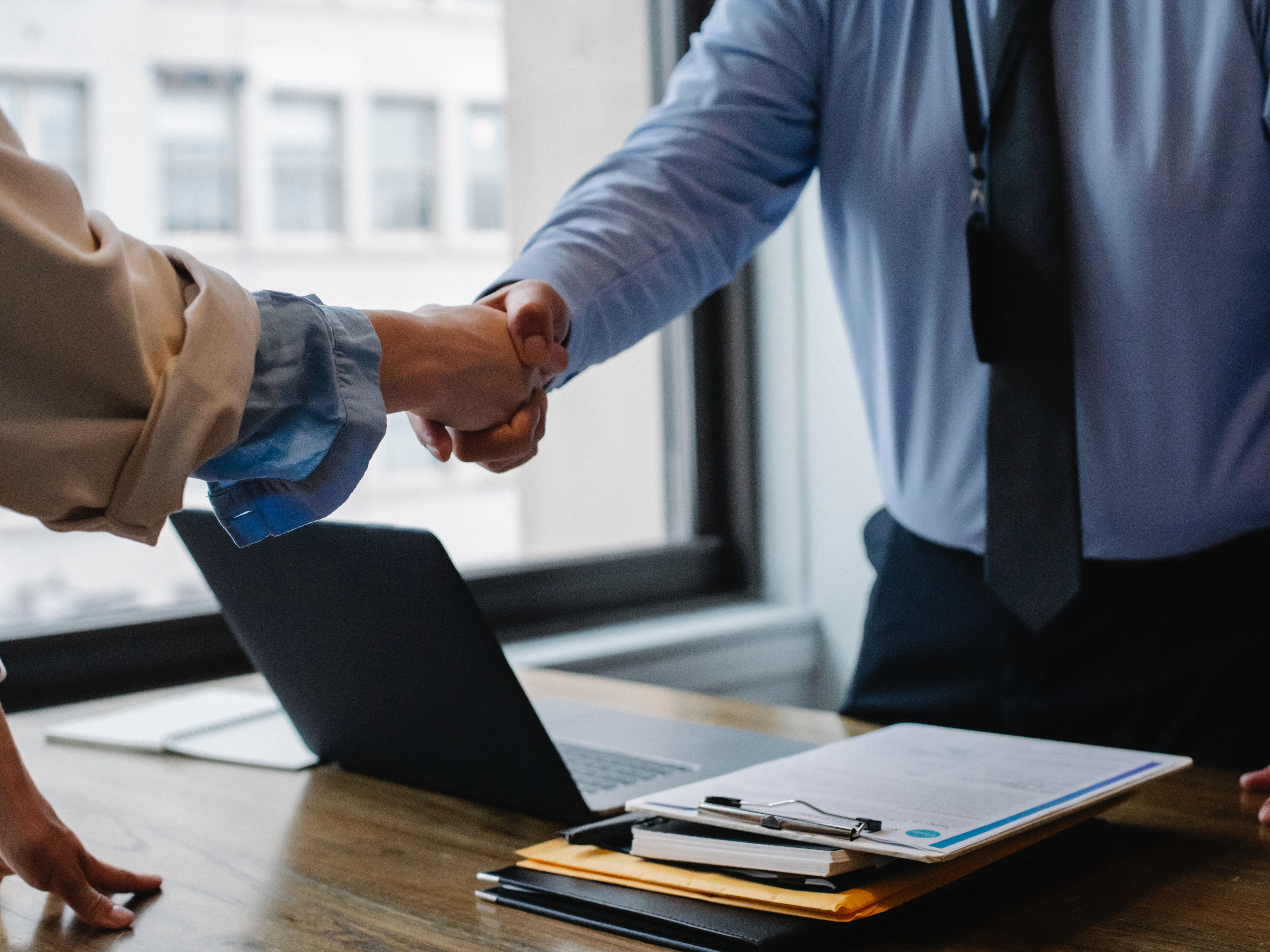 Two people wearing business attire shake hands over a laptop and a clipboard, near a window.