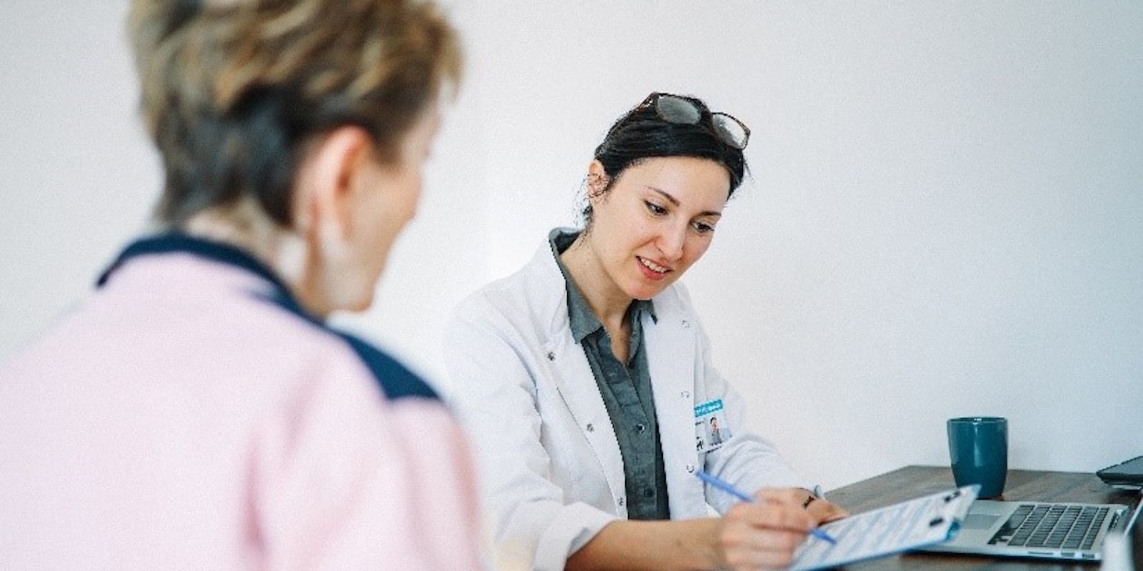 A female doctor in a white coat is sitting at a desk, smiling while writing on a clipboard. She is engaging with an older female patient, who is wearing a pink and navy sweater. A laptop, documents, and a blue mug are on the desk in a medical office setting.