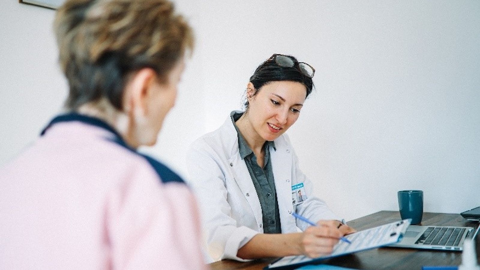 A female doctor in a white coat is sitting at a desk, smiling while writing on a clipboard. She is engaging with an older female patient, who is wearing a pink and navy sweater. A laptop, documents, and a blue mug are on the desk in a medical office setting.