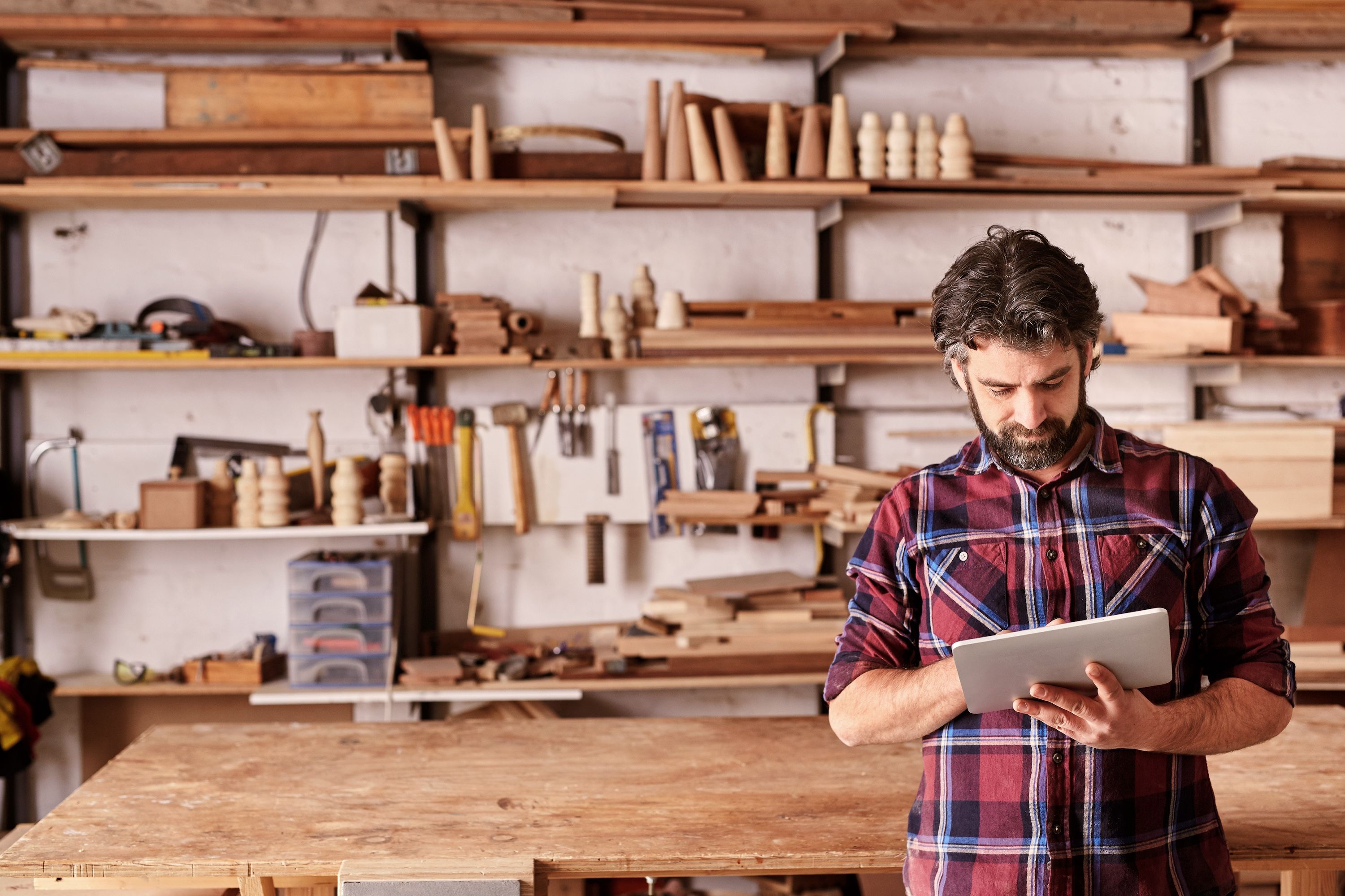 A carpenter in a plaid shirt does business on an iPad in his shop.