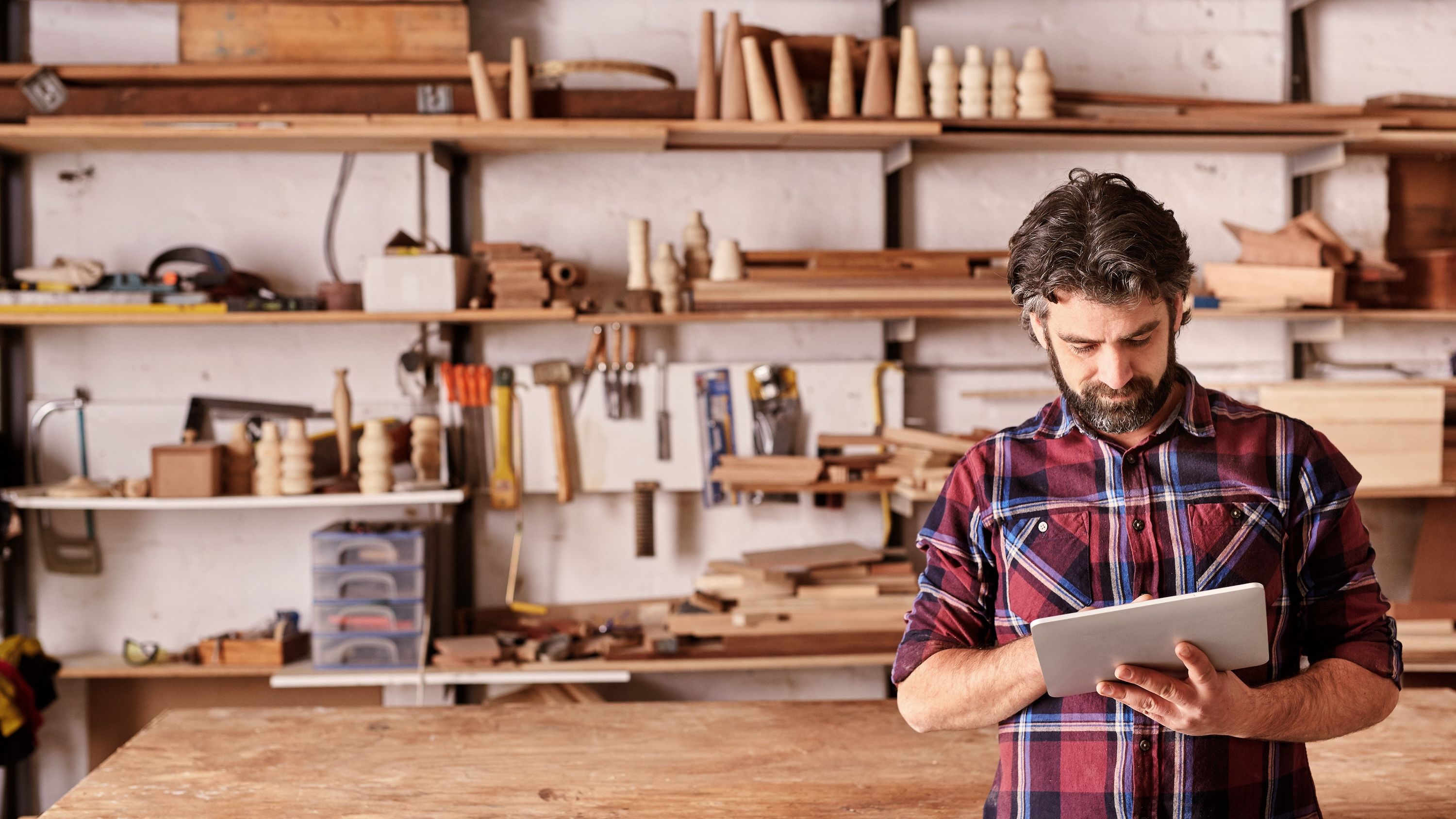 A carpenter in a plaid shirt does business on an iPad in his shop.