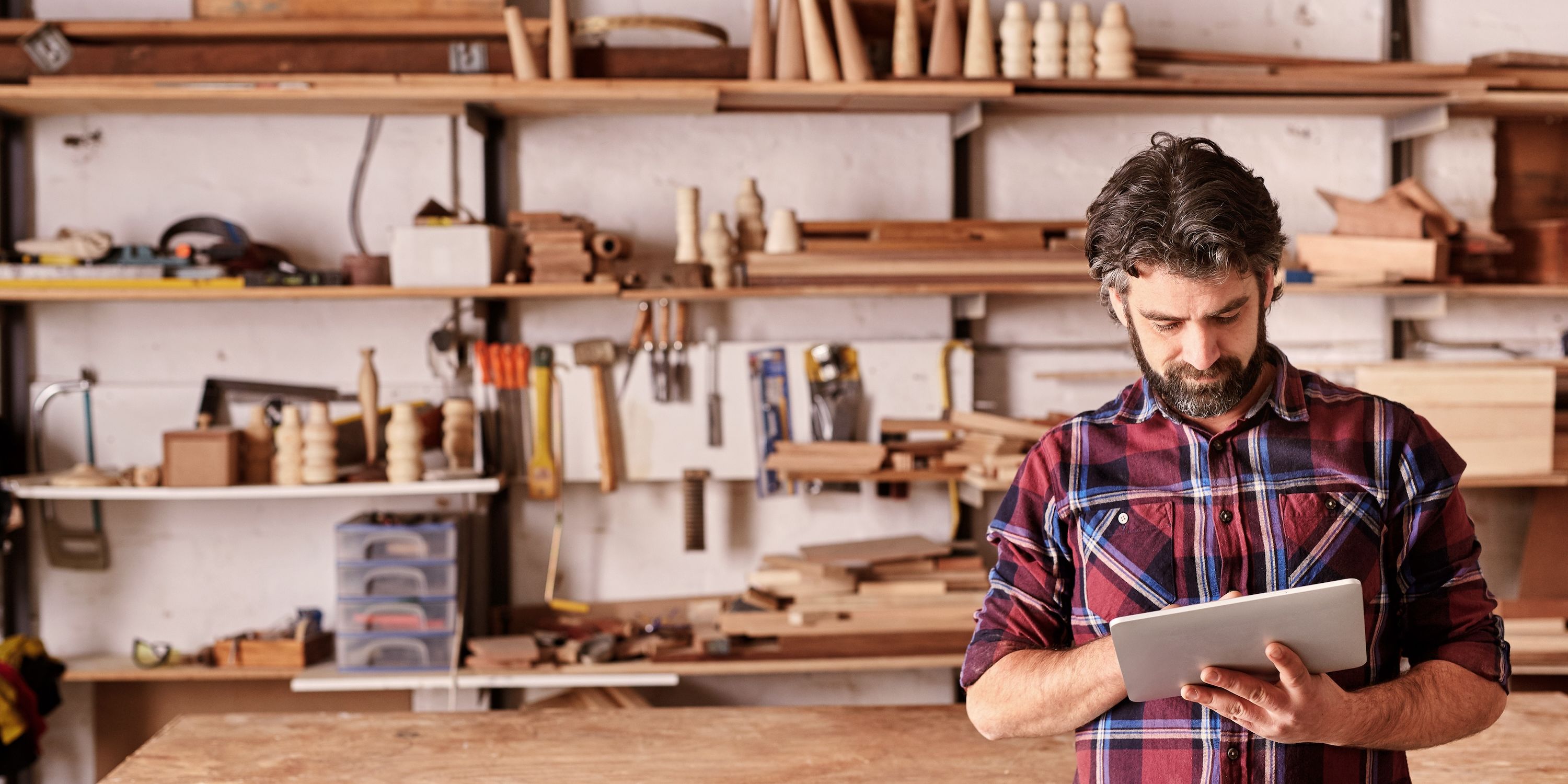 A carpenter in a plaid shirt does business on an iPad in his shop.
