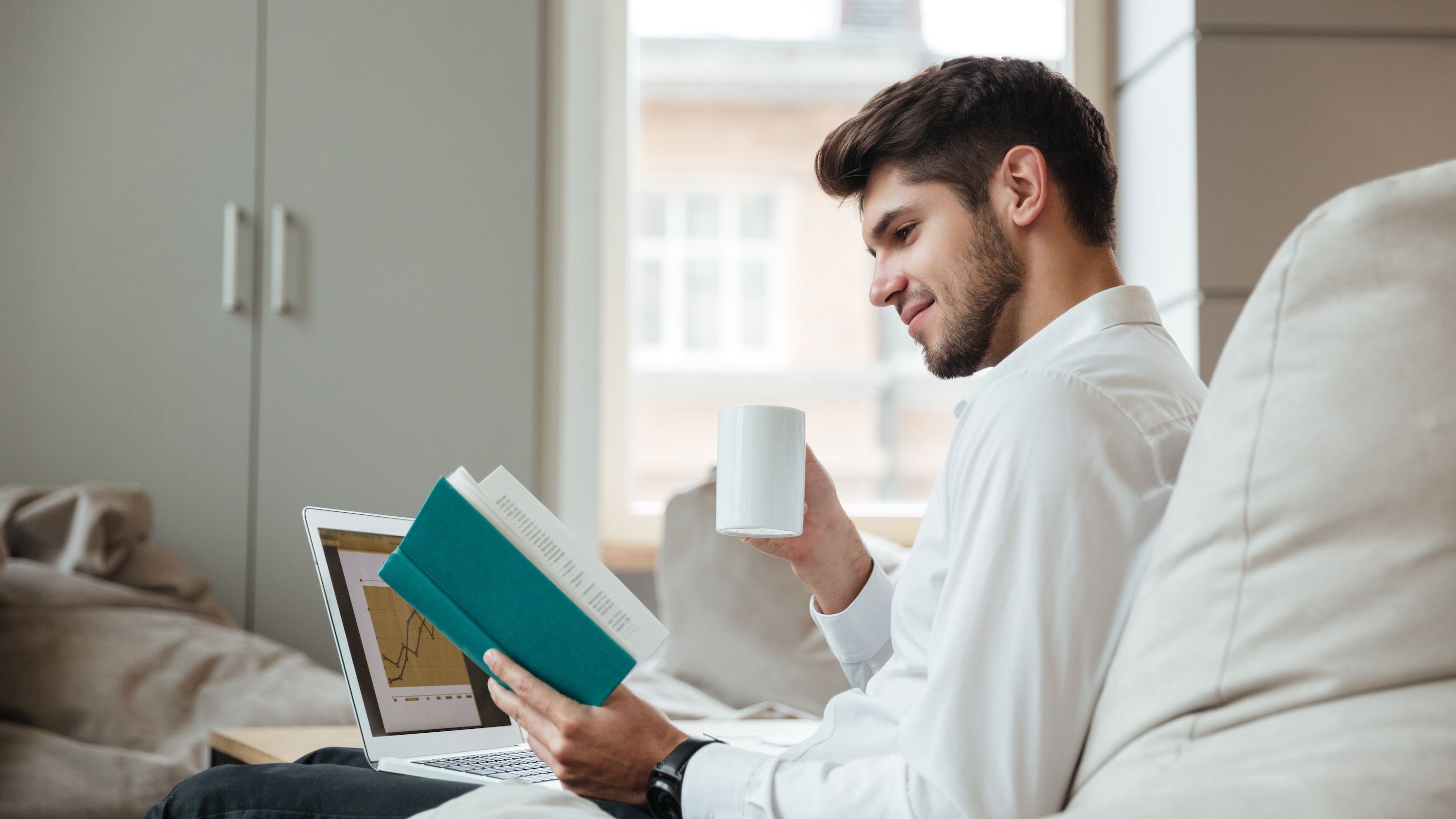 An aspiring young businessman reads business books while looking at line graphs of numbers going up on his laptop.