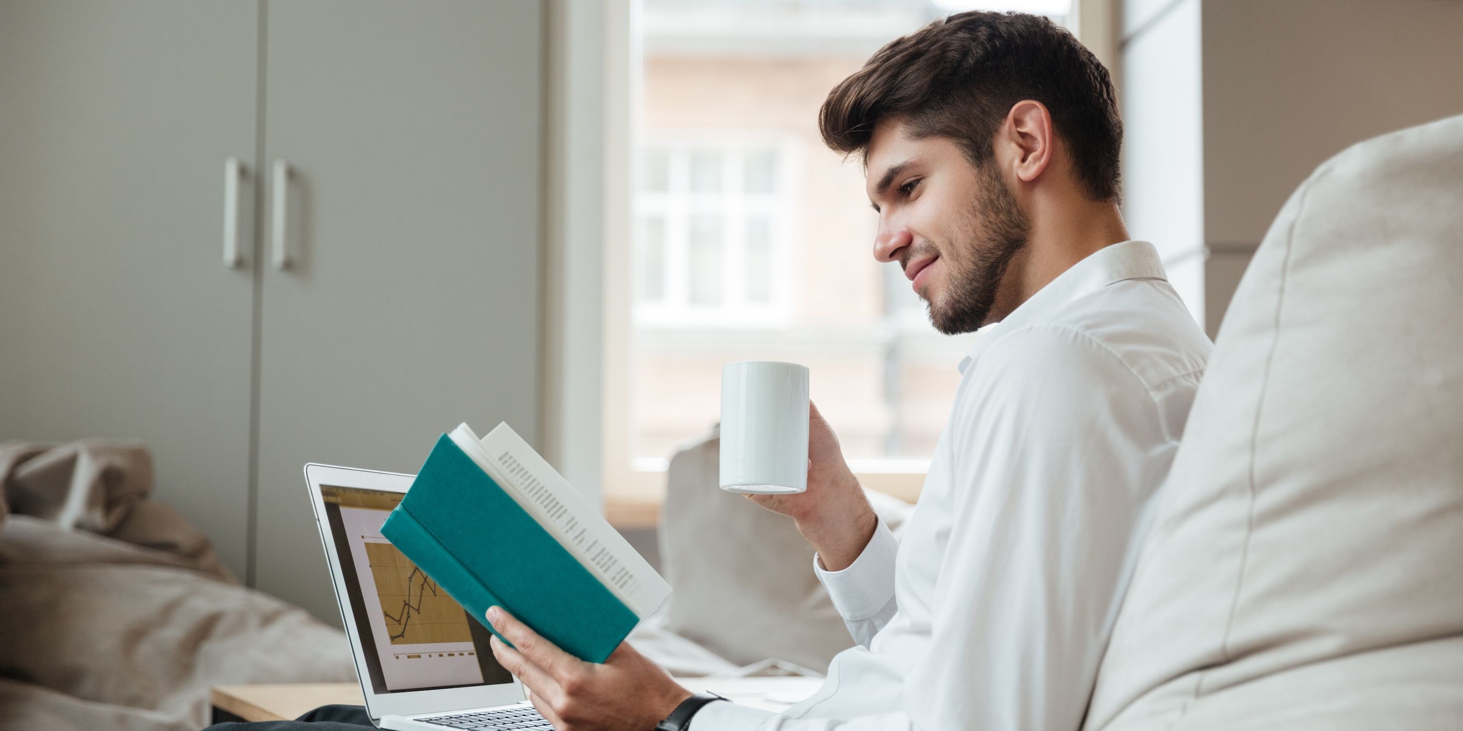 An aspiring young businessman reads business books while looking at line graphs of numbers going up on his laptop.