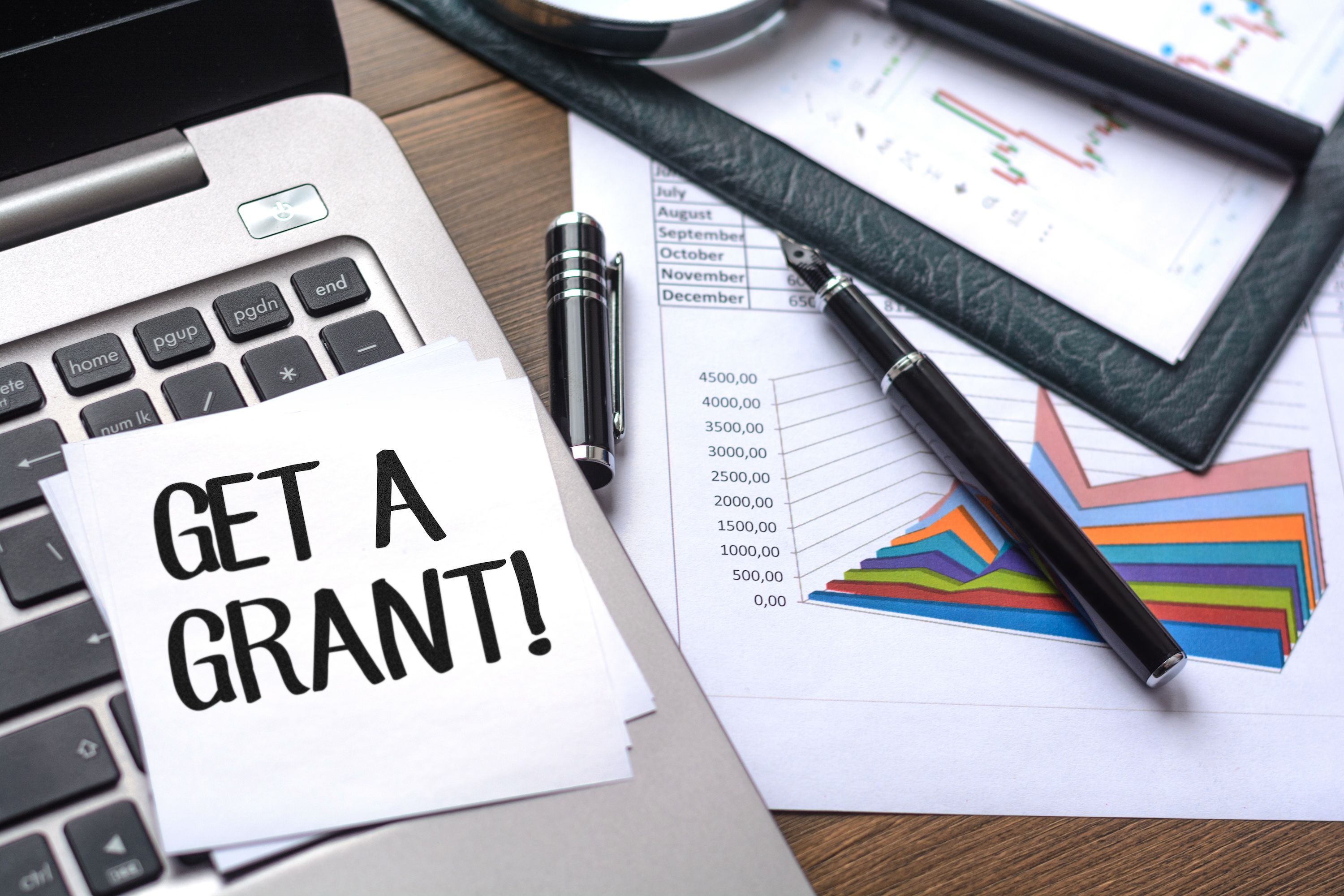 A stack of budget graphs and documents next to a sticky note labeled 'Get A Grant!'