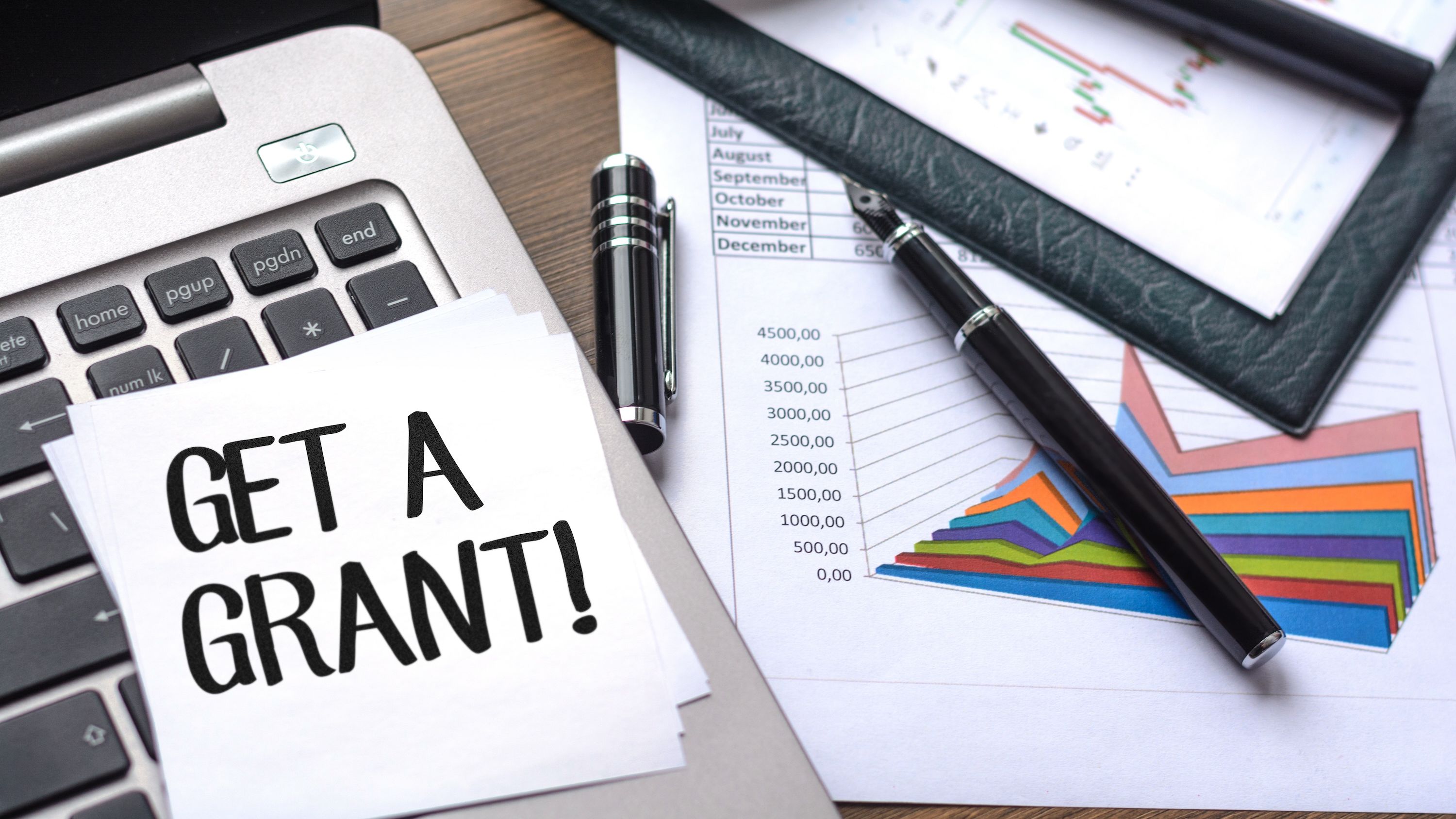 A stack of budget graphs and documents next to a sticky note labeled 'Get A Grant!'