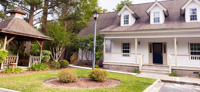 A cute two story office with porch on right with lamppost in front and a gazebo on left with pine trees in the background