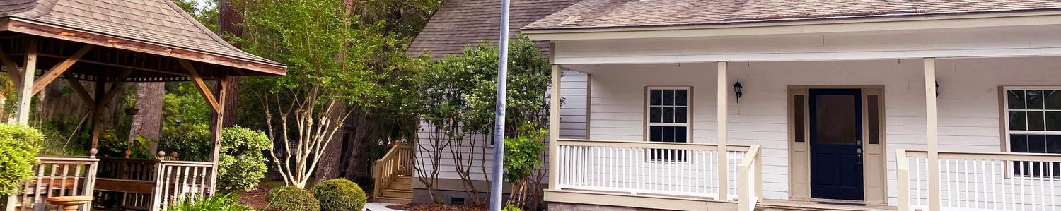 A cute two story office with porch on right with lamppost in front and a gazebo on left with pine trees in the background