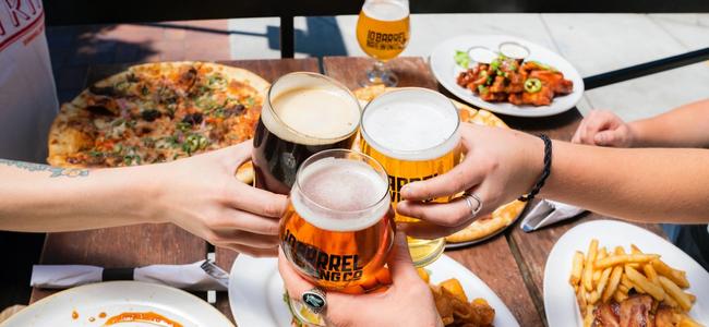 Overhead of three friends touching beer glasses over a table with many different plates of food.