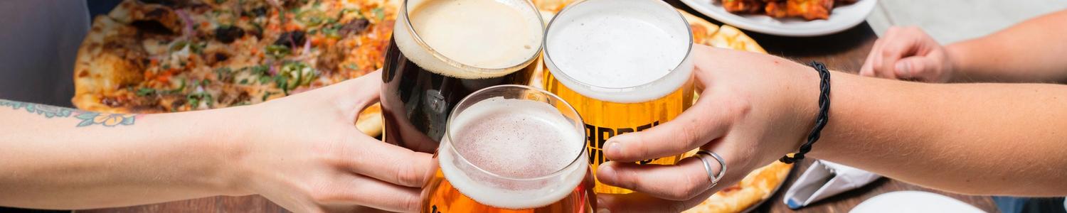 Overhead of three friends touching beer glasses over a table with many different plates of food.