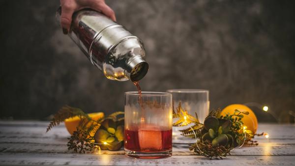 A cocktail being poured from a stainless steel shaker over a large rock. Surrounded by two lemons, fern sprouts, and succulents. Painted cement background.