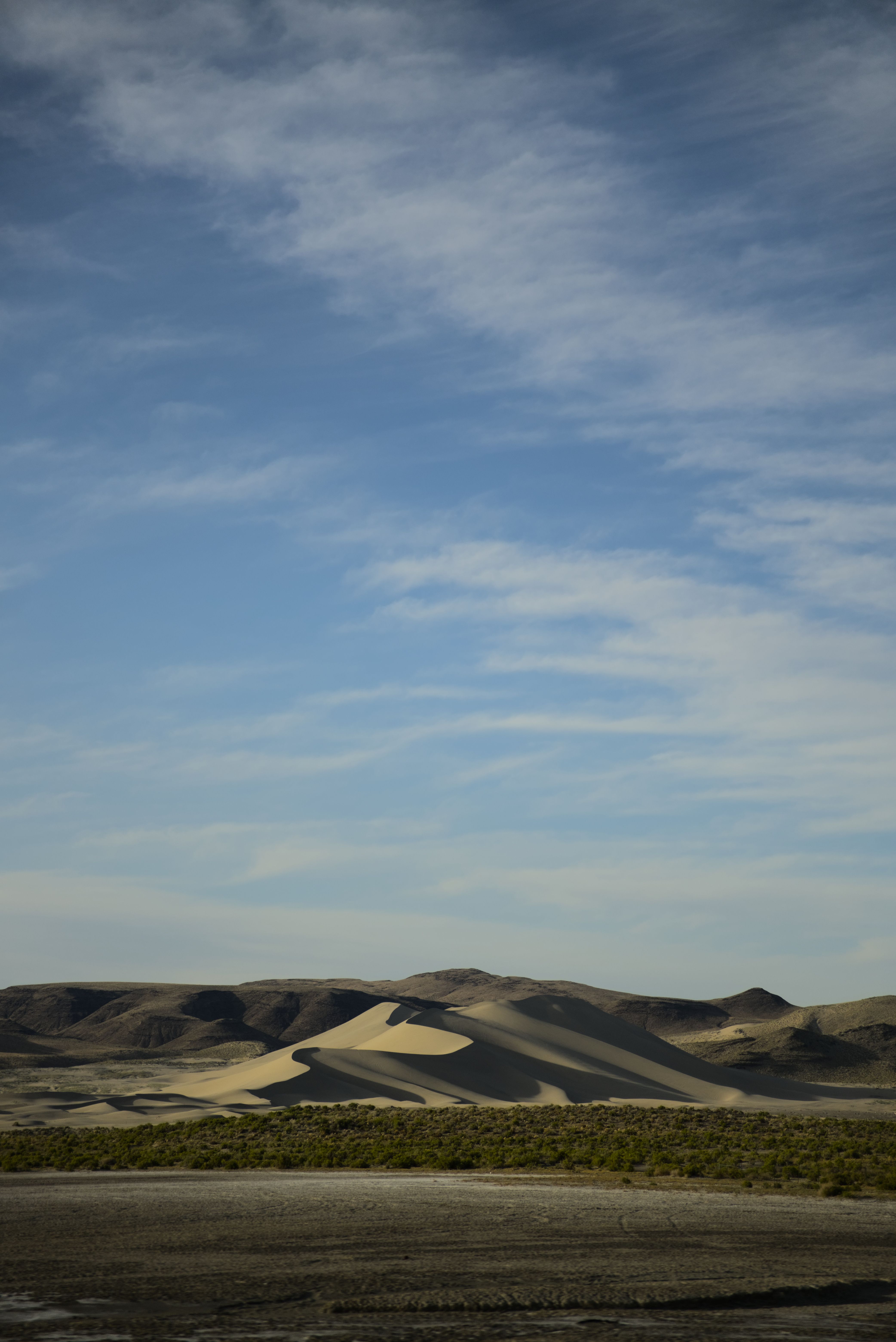 A sand dune at sunset, shadowed on the right side and lit on the left, under a blue sky with whispy clouds.