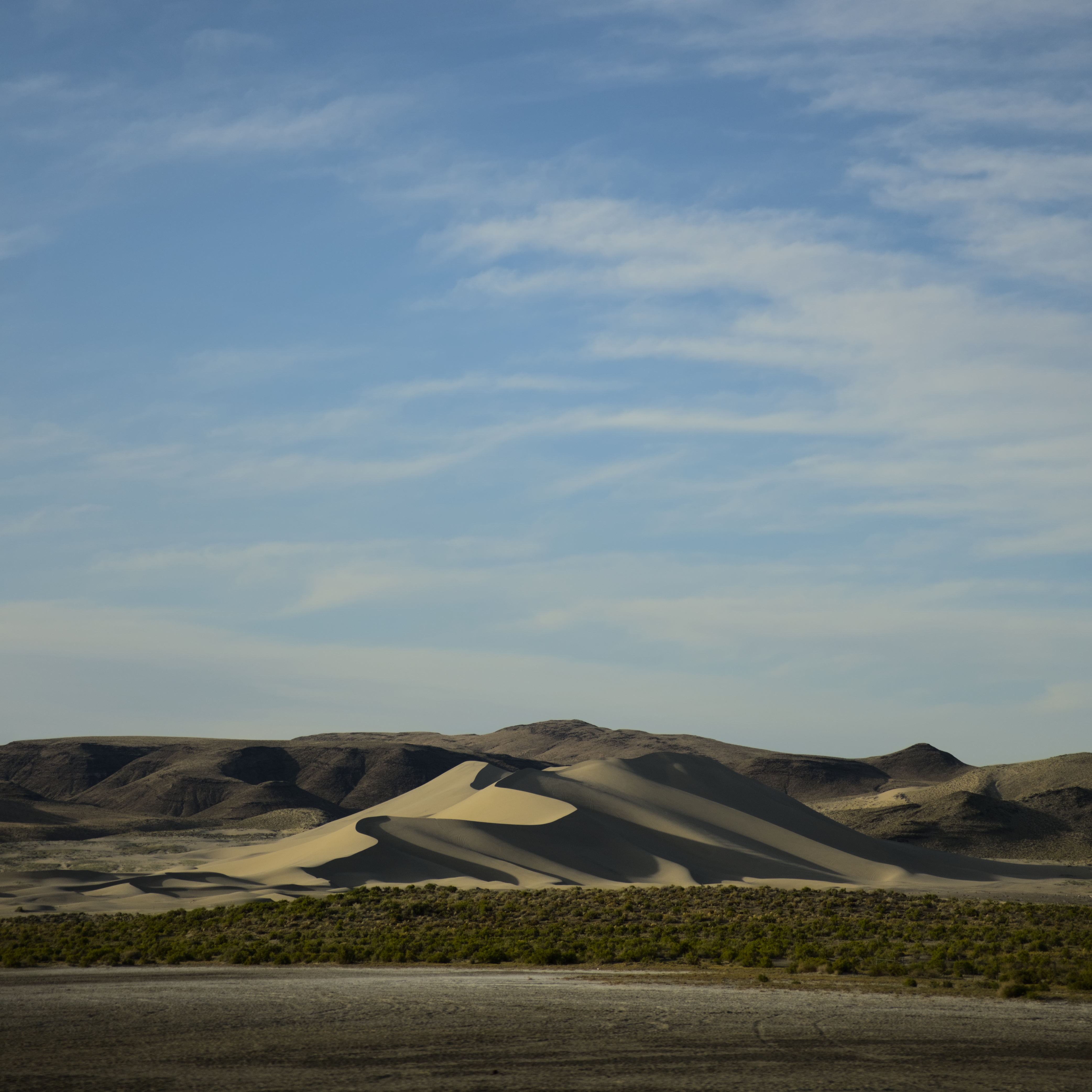 A sand dune at sunset, shadowed on the right side and lit on the left, under a blue sky with whispy clouds.