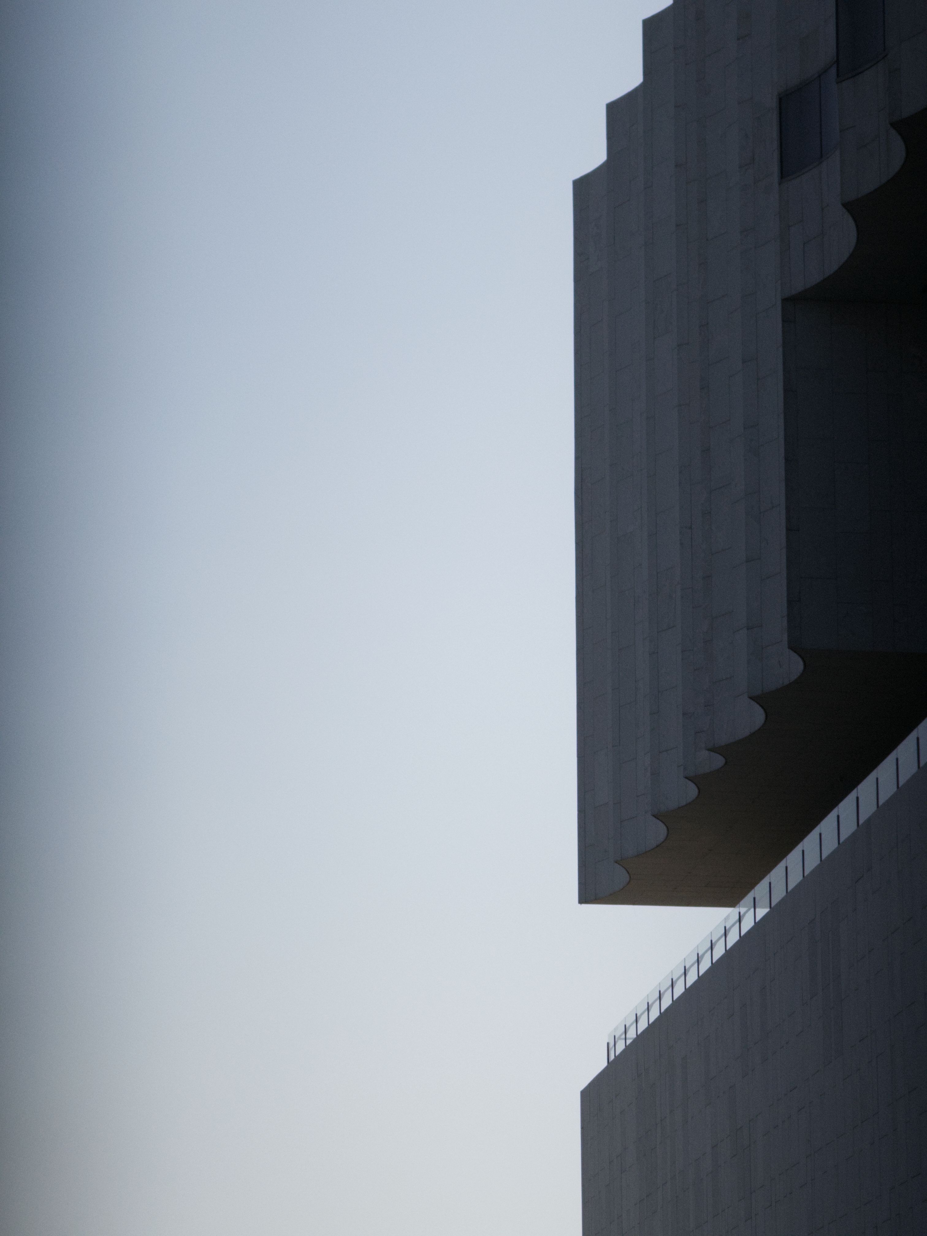 The scalloped corner of a large concrete building with a balcony, silhouetted against the sky
