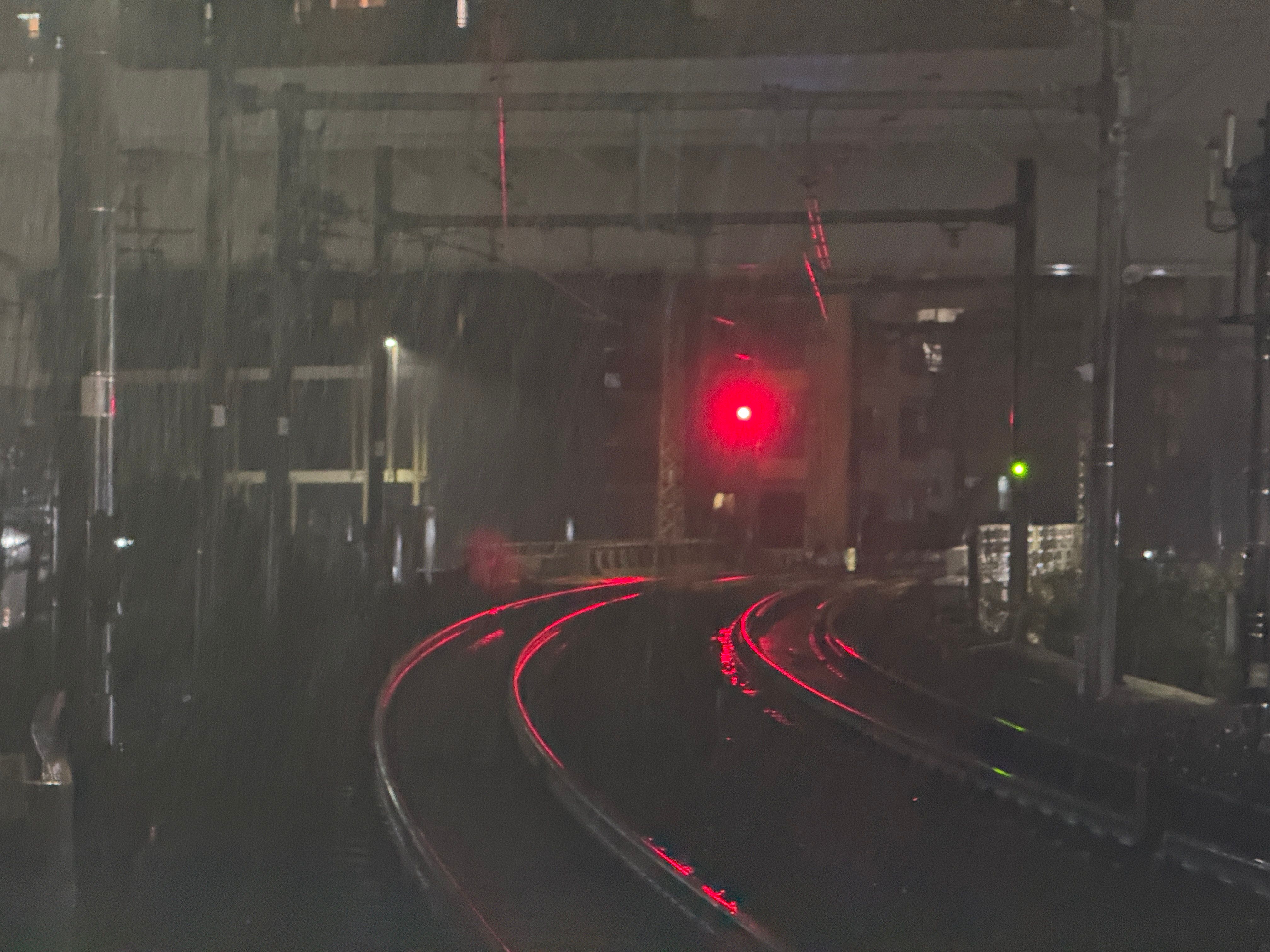 Two sets of curving railroad tracks in the rain at night. A single red light illuminates them and reflects off the tracks.