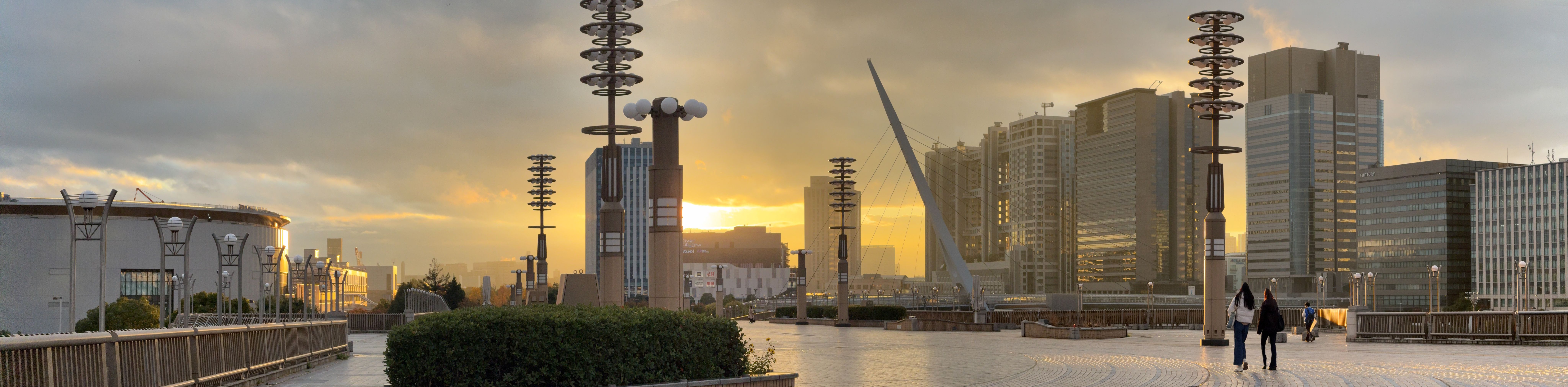 A panorama of a sunset over a long pedestrian walkway with some tall buildings in the background