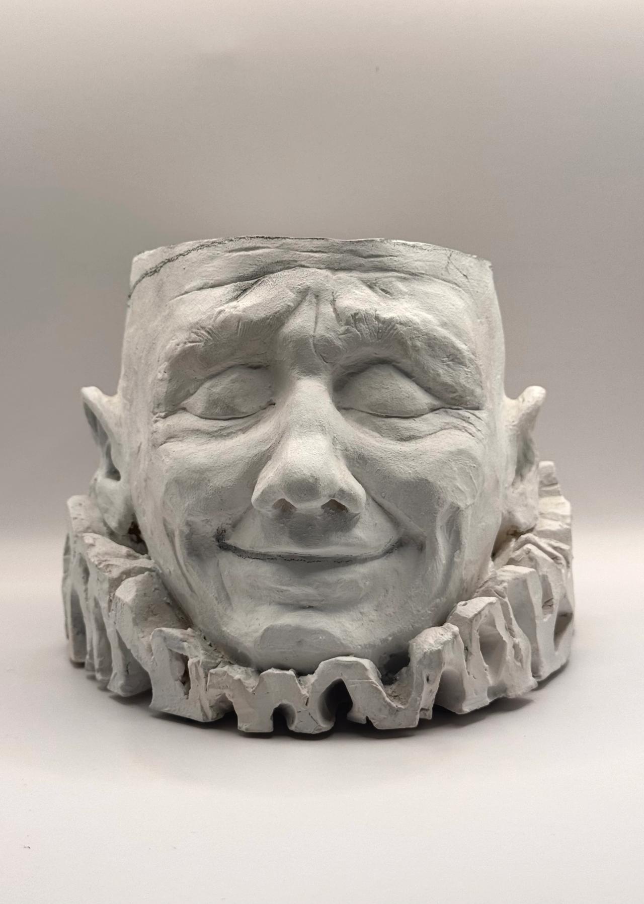 Clay sculpture of a smiling face with closed eyes and a ruffled collar, front view