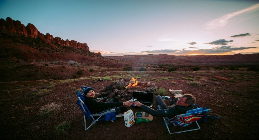 Two campers toasting drinks by a campfire at sunset in southern Utah's red rock desert