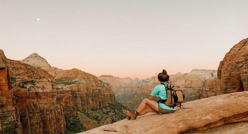 A female hiker with a backpack sits on a sandstone ledge at Zion National Park's Overlook Point Trail