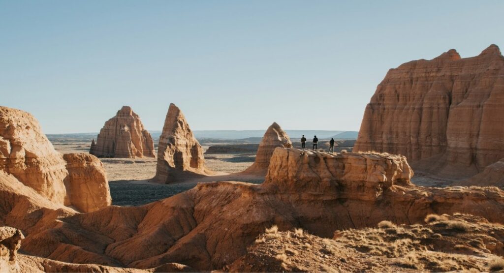 Three hikers stand atop a sandstone ridge surrounded by towering cathedral-like rock spires in Capitol Reef National Park's Cathedral Valley