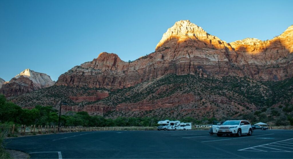 An RV and vehicles parked in the visitor center lot at Zion National Park, with sunlit red and white sandstone peaks