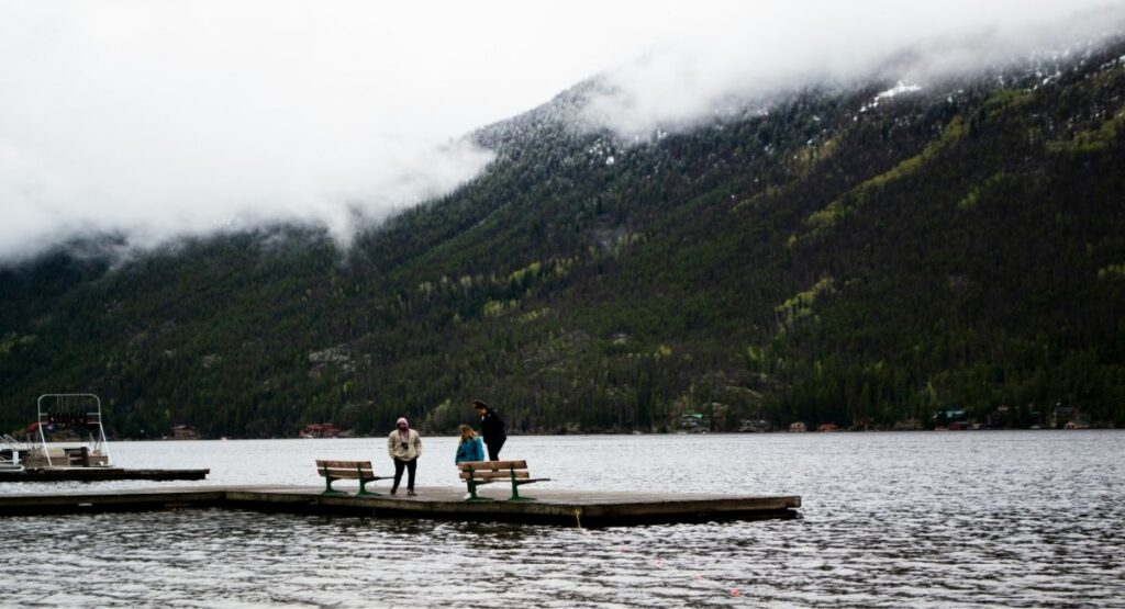 Three visitors on a lakeside dock at Grand Lake, Colorado, with a forested mountain dusted in snow and low clouds 