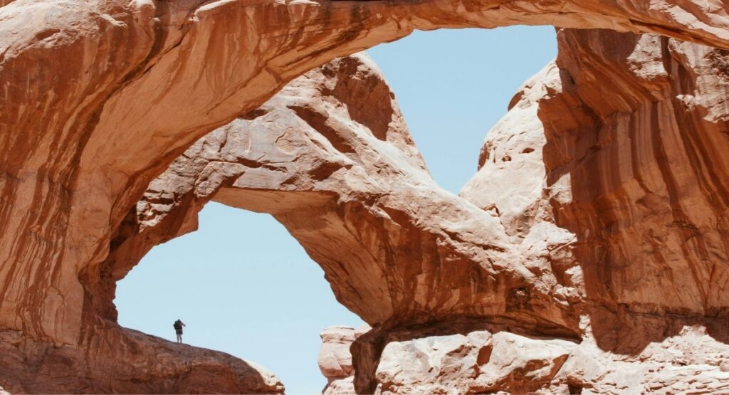 A lone hiker stands dwarfed beneath the massive twin spans of Double Arch at Arches National Park
