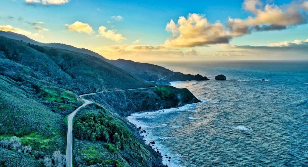 Aerial view of Highway 1 winding along the rugged Big Sur coastline, with forested cliffs dropping to rocky shores