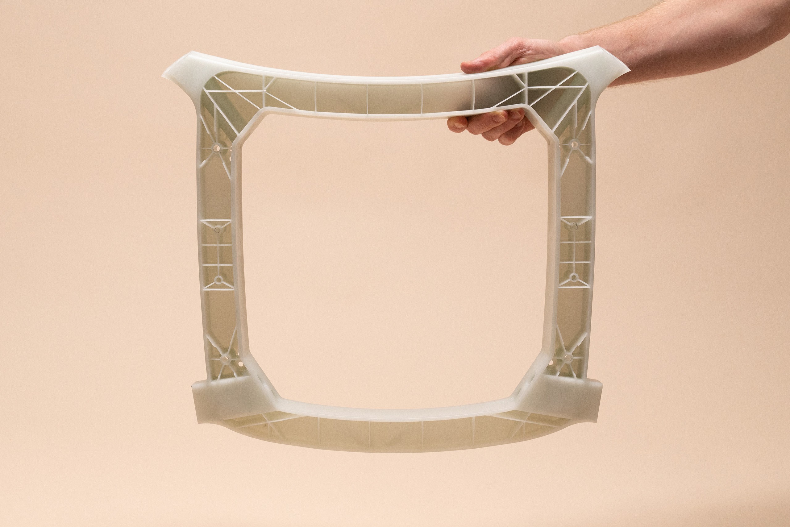 A hand is holding a white translucent plastic component in the form of a ring-shaped square. In the background there is a pale rose colored wall. 