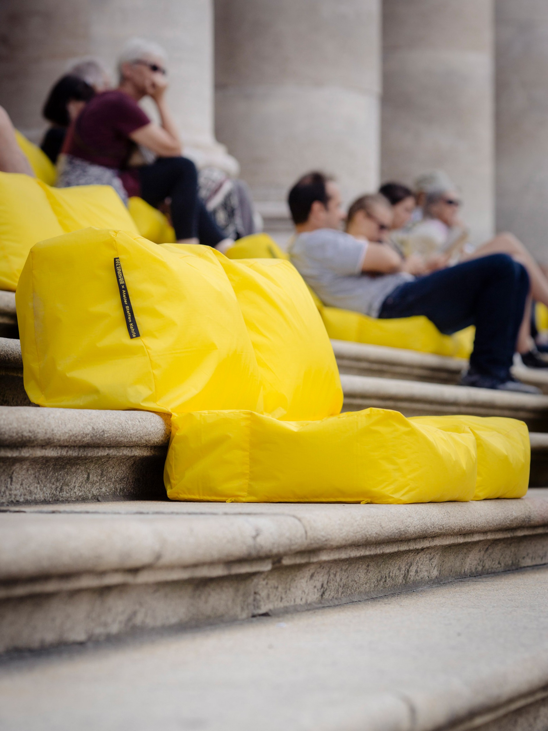 A bright yellow bean bag seat placed on the stone steps of a classical building, with large stone columns visible in the background. The bean bag is shaped to form a low seat with a backrest, made from a shiny, lightweight fabric. A small label is visible on the side. The vivid yellow contrasts boldly with the grey stone surroundings.